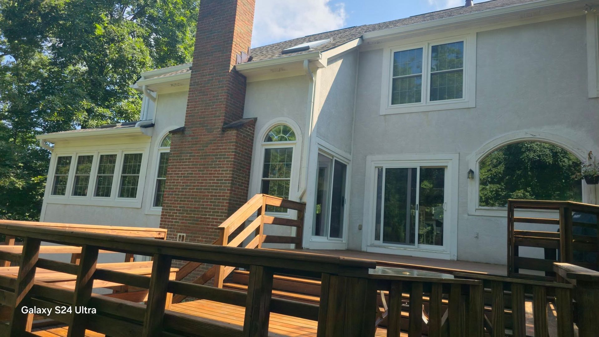 Back of a two-story house with a deck. Brick chimney, stucco walls, windows. Brown railing in foreground. Sunny day.