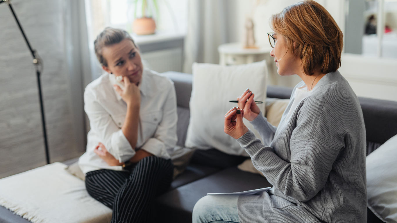 Woman in therapy session with therapist on sofa. One gestures, other looks concerned.