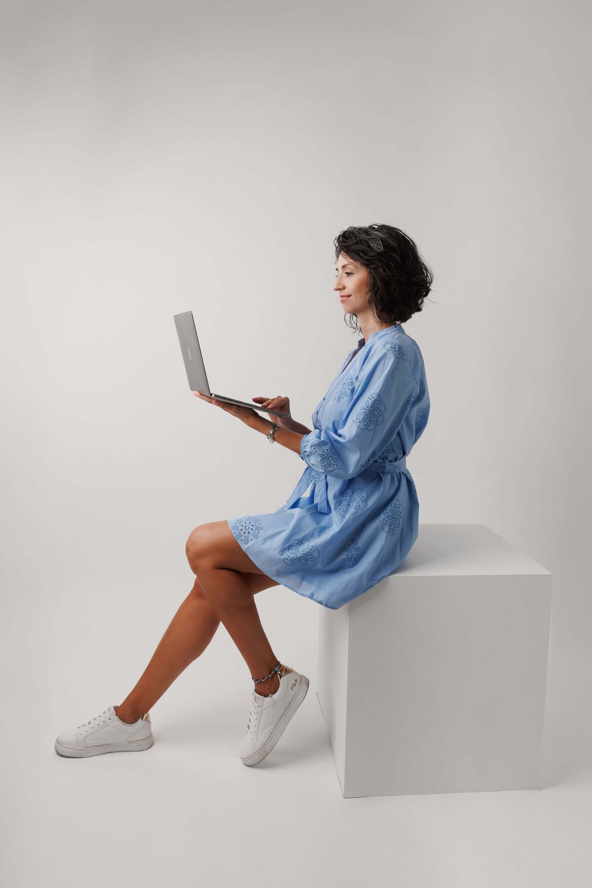 Una mujer con un vestido azul está sentada en un cubo blanco usando una computadora portátil.