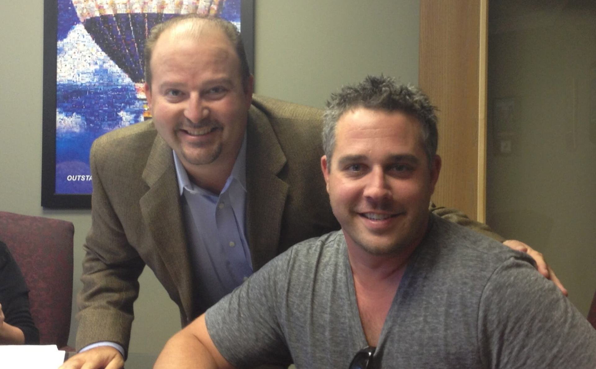 Two men are posing for a picture in front of a picture of a hot air balloon.