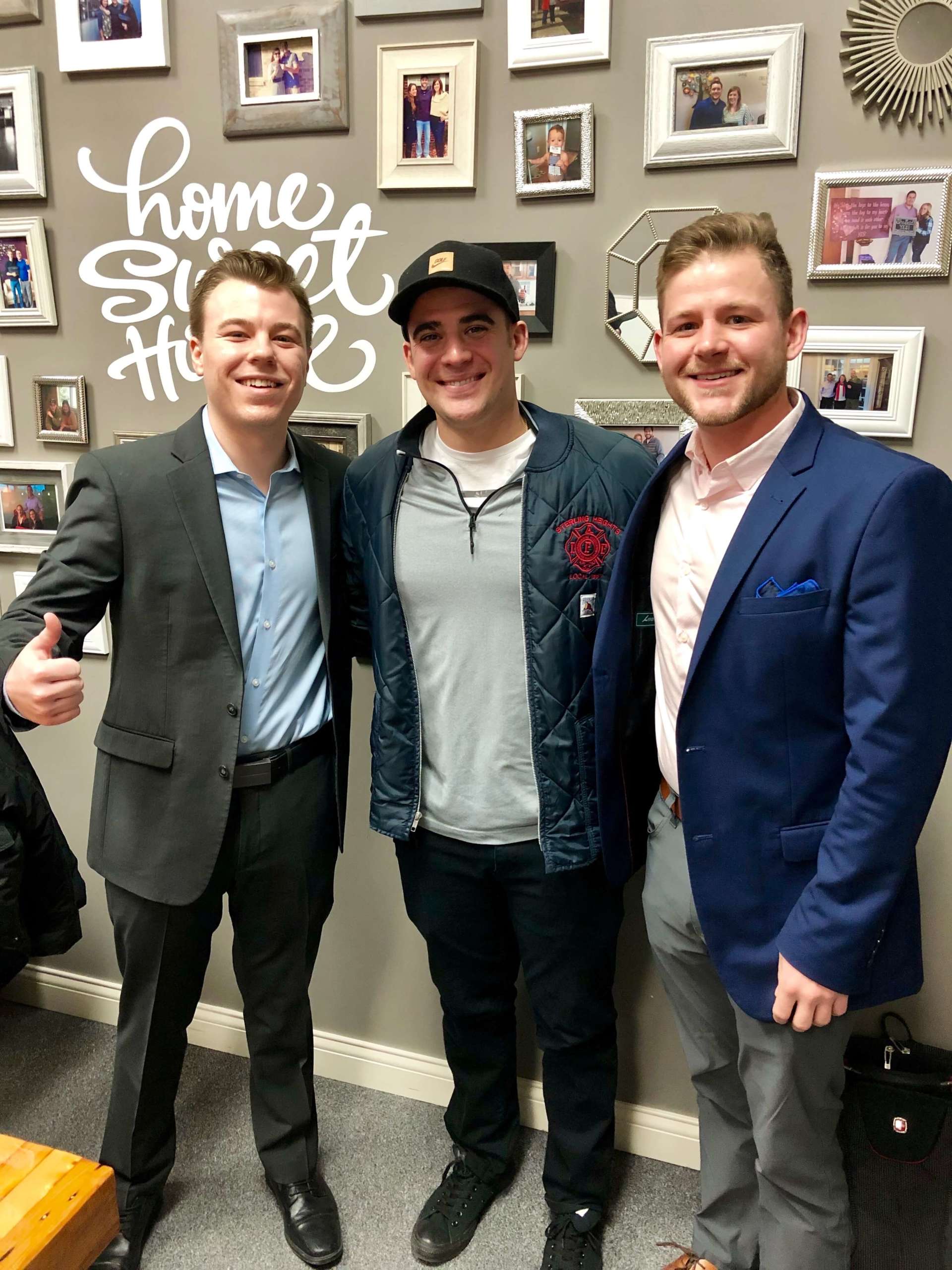 Three men are posing for a picture in front of a wall that says home sweet home.