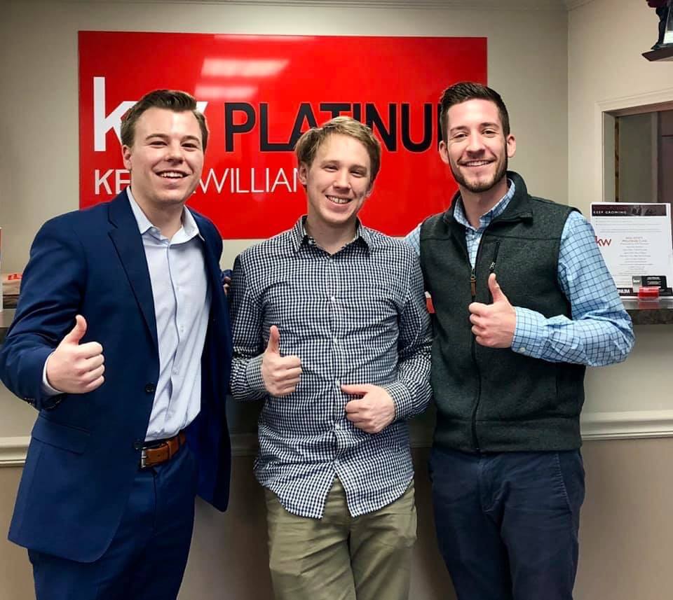 Three men are posing for a picture in front of a sign that says kw williams.