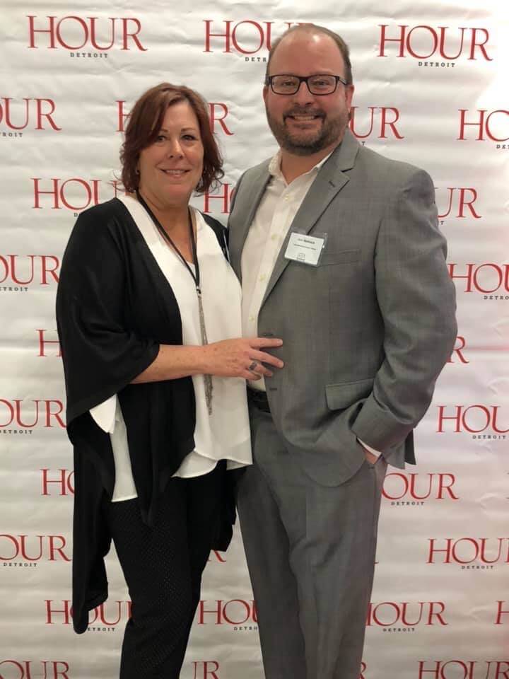 A man and a woman are posing for a picture in front of a wall that says hour.