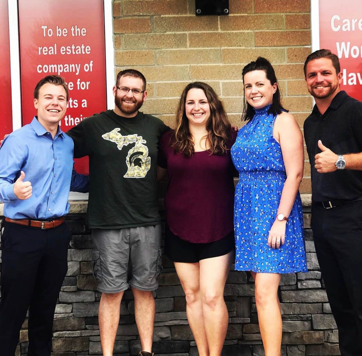 A group of people standing in front of a sign that says to be the real estate company.