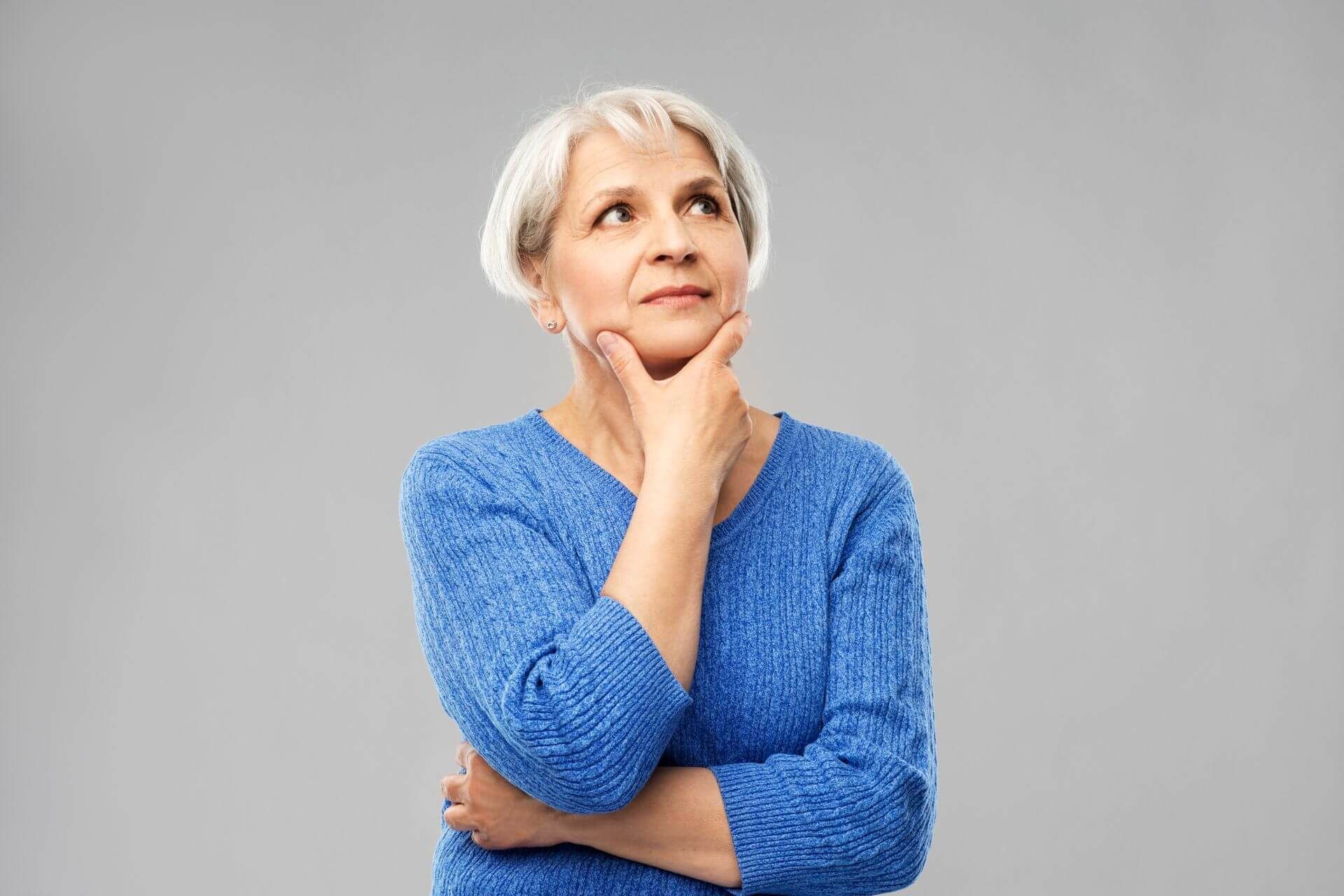 Woman with short white hair, blue sweater, looking up with a thoughtful expression, hand on chin.