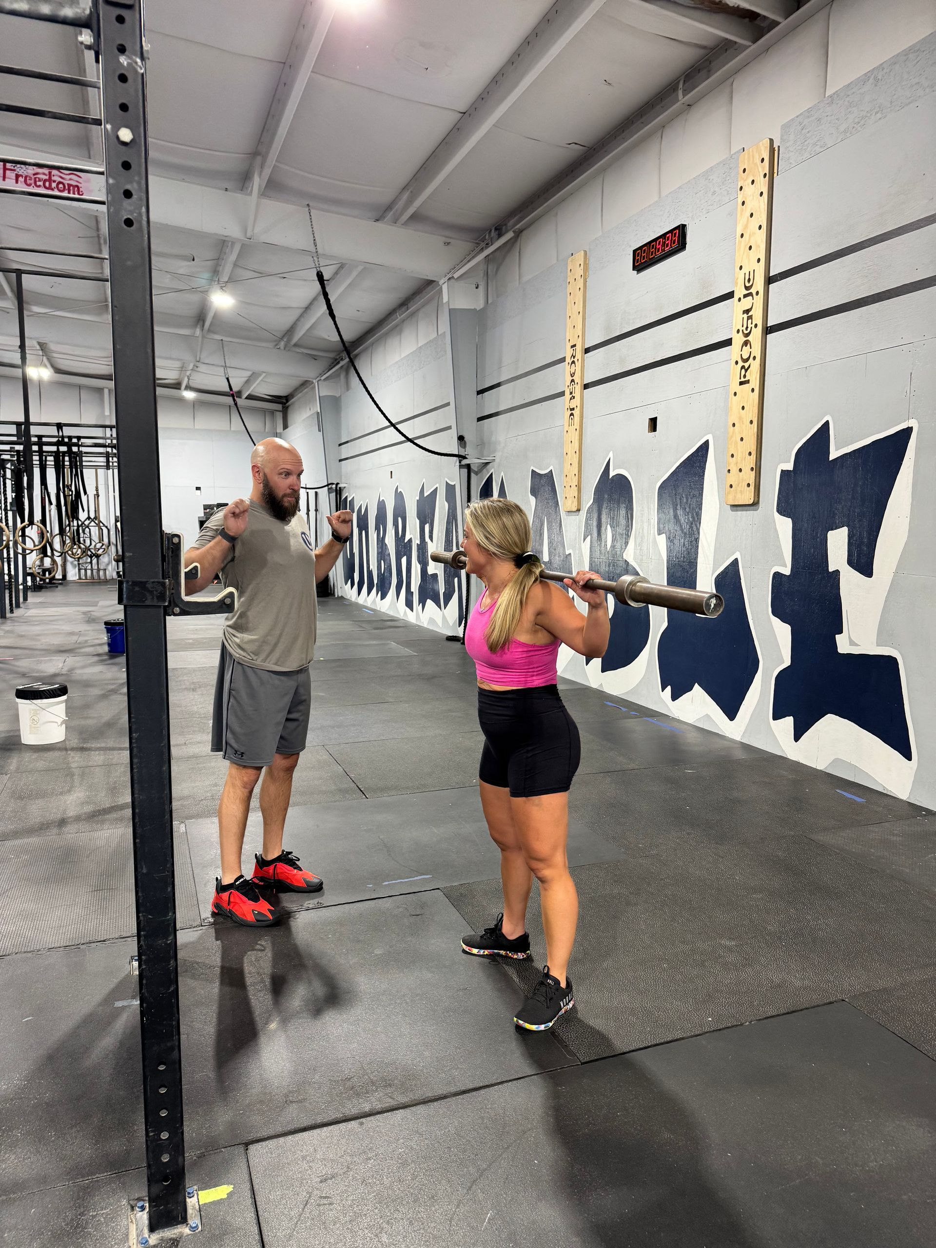 Two people exercising in a CrossFit gym, facing each other with arms raised beside a rig and wall markings.