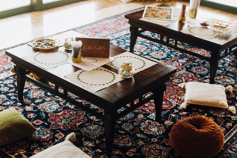 Two low, wooden coffee tables arranged on a patterned rug with floor cushions, plates, and a sign reading