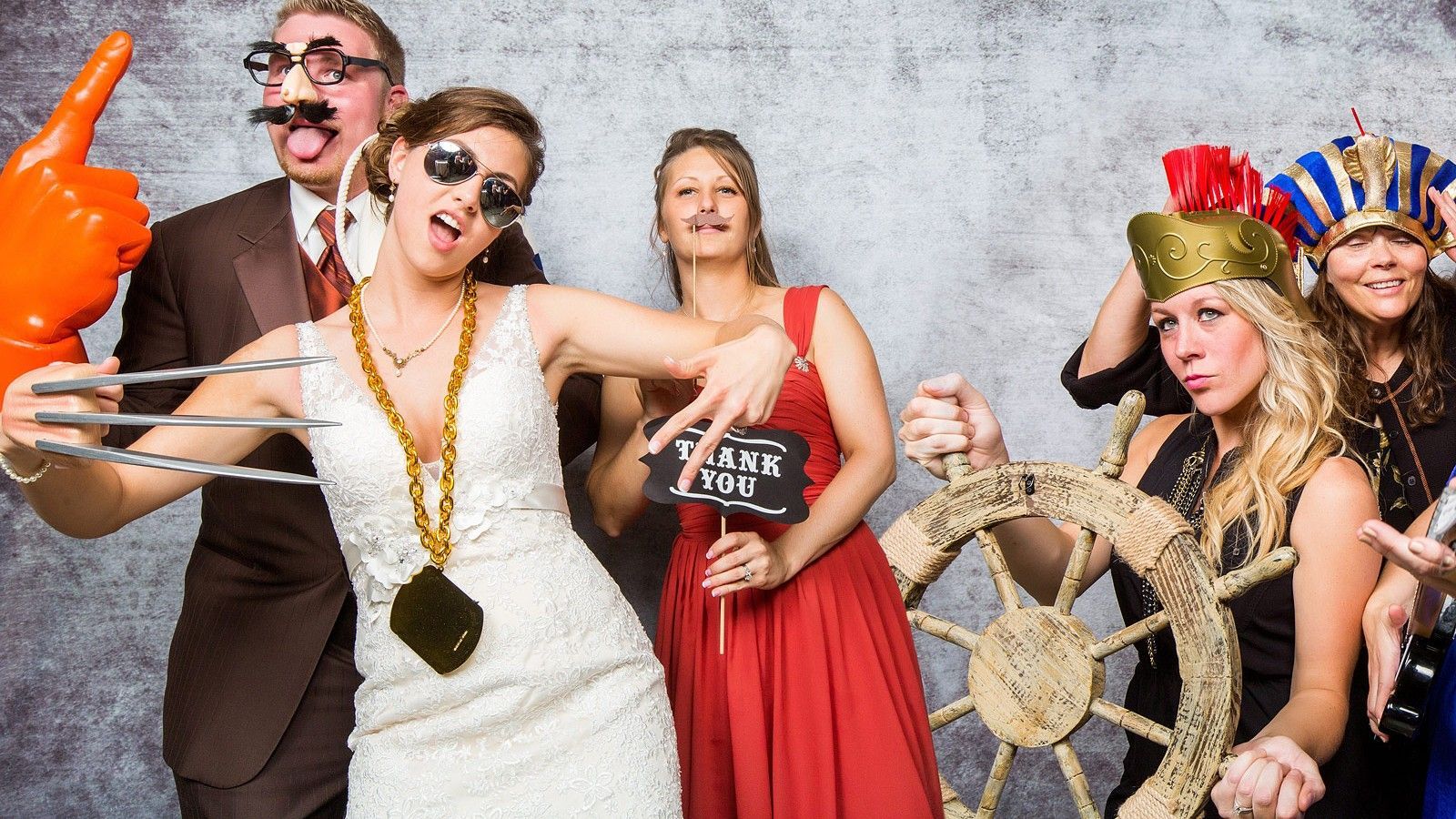 Vijf mensen in kostuums en met fotohokjesaccessoires poseren tegen een grijze achtergrond, sommigen met props