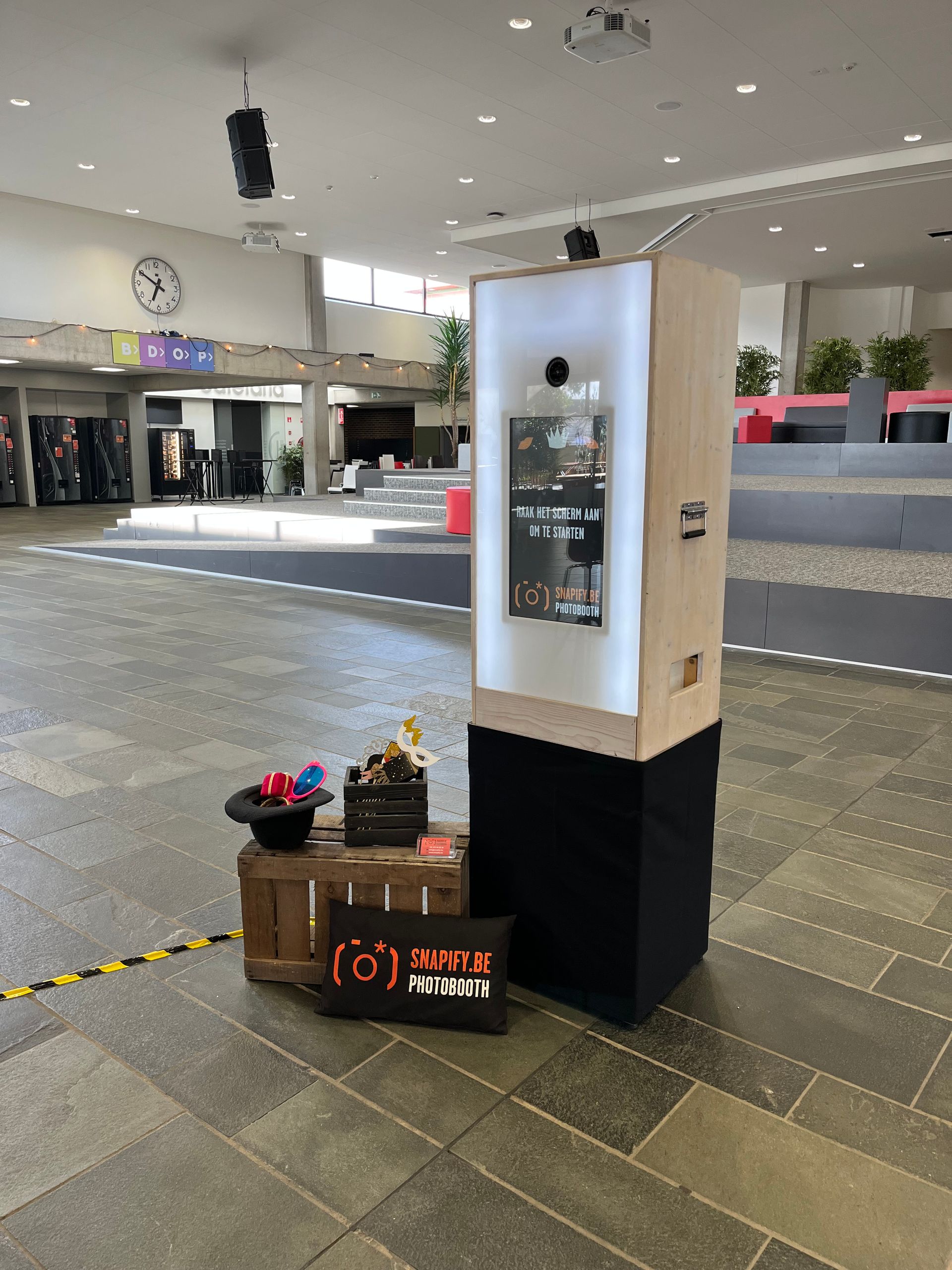 In een ruime hal staat een houten fotocabine met props en een reclamebord met het bedrijfslogo aan de voet.
