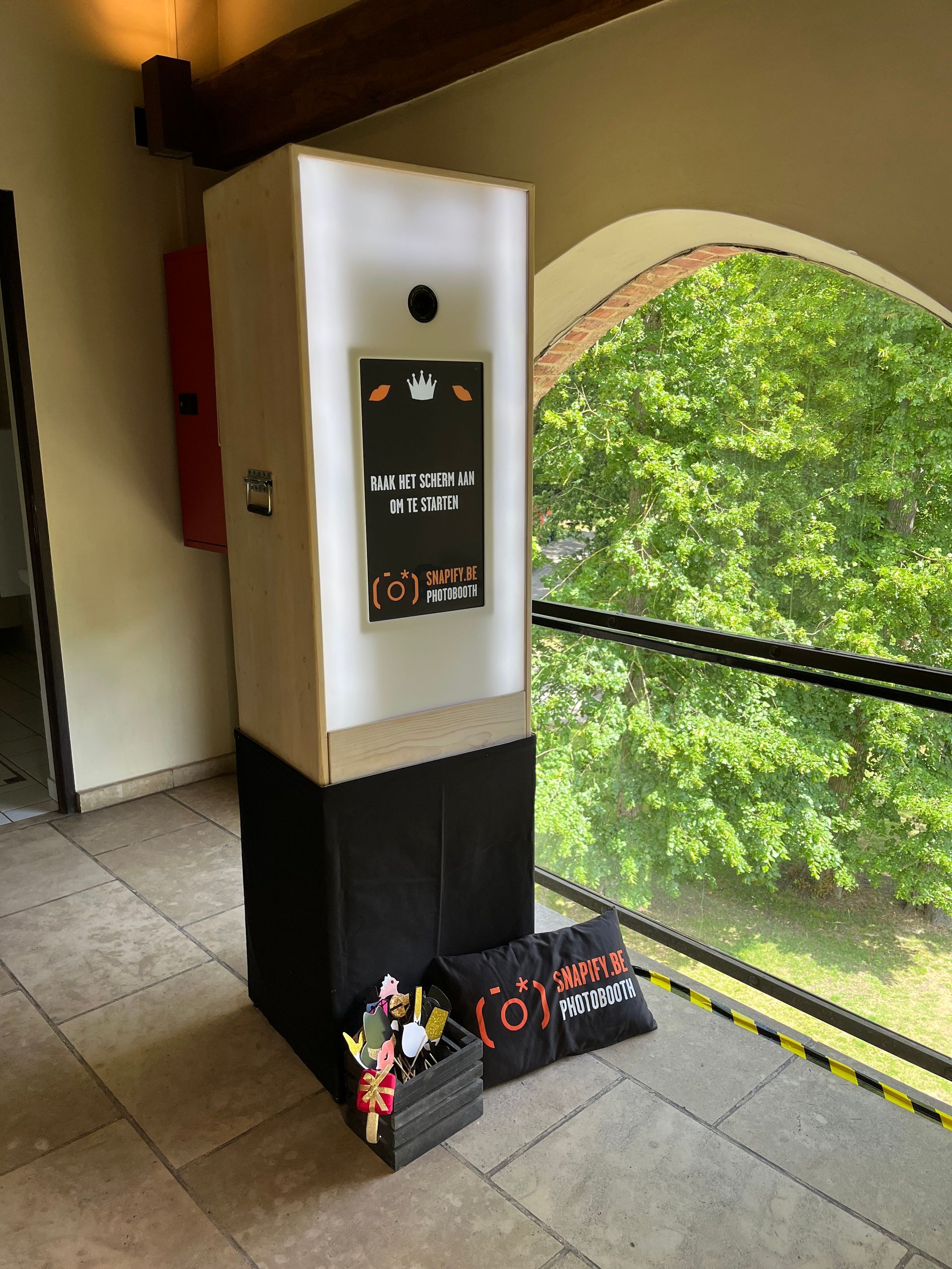 Een photobooth staat op een tegelvloer vlakbij een groot raam, met een zwarte kist met rekwisieten en een bord aan de voet.