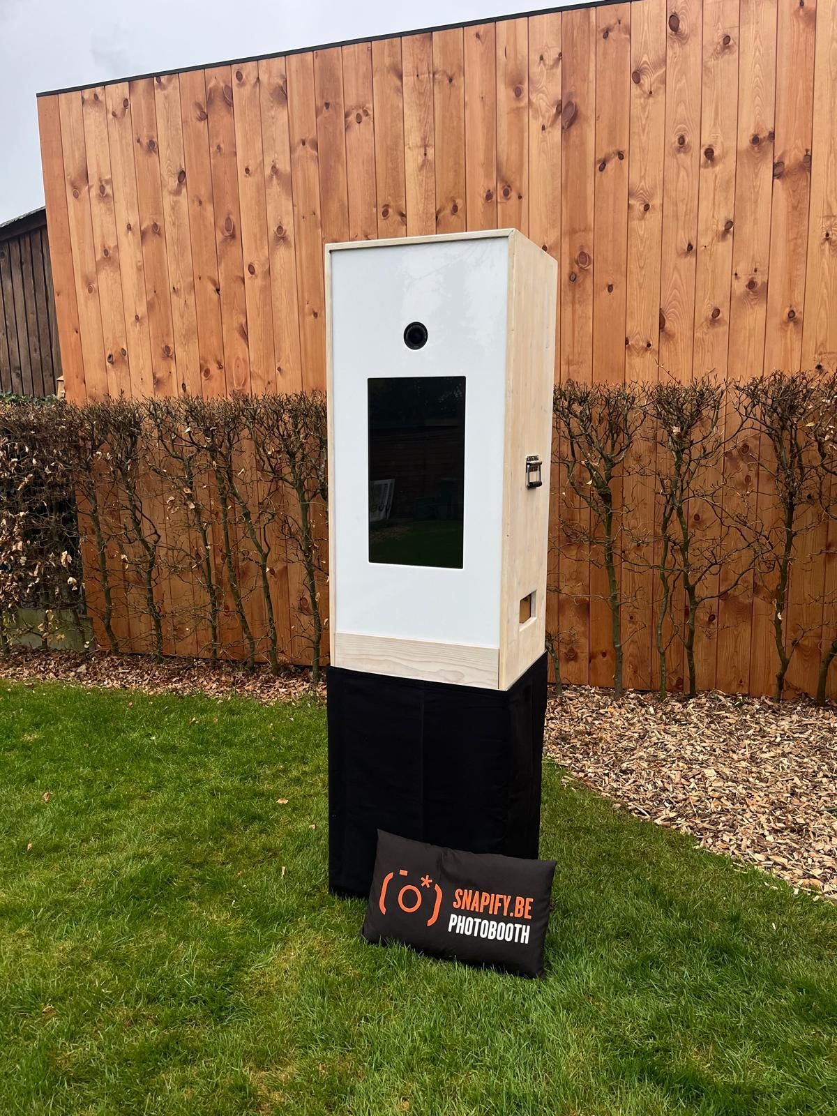 Een verplaatsbare houten fotocabine met scherm en camera, die op het gras voor een houten hek staat.