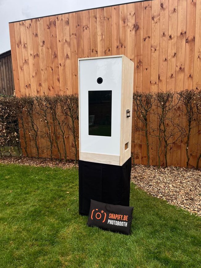Een houten photobooth staat op een zwarte sokkel op een gazon voor een houten hek, met een reclamebord ernaast.