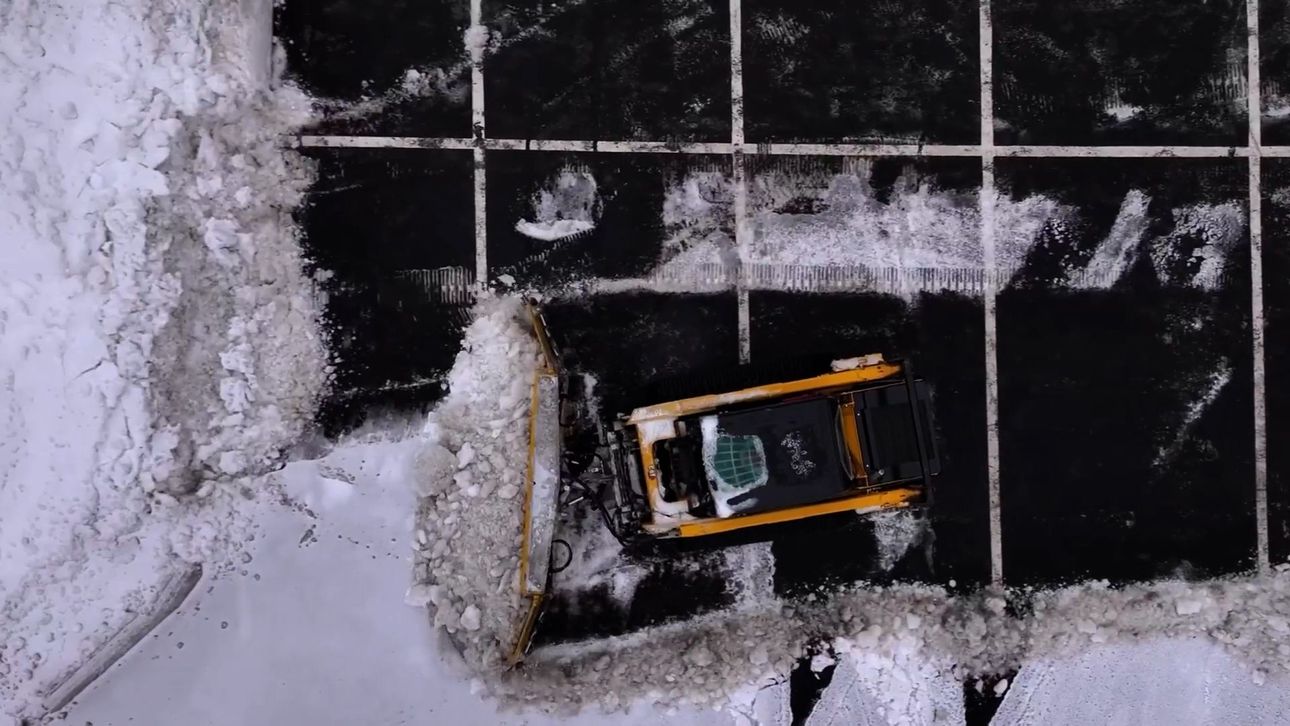 Park with green grass, trees, and a red sports area. A person sits on a bench. Buildings in the background.