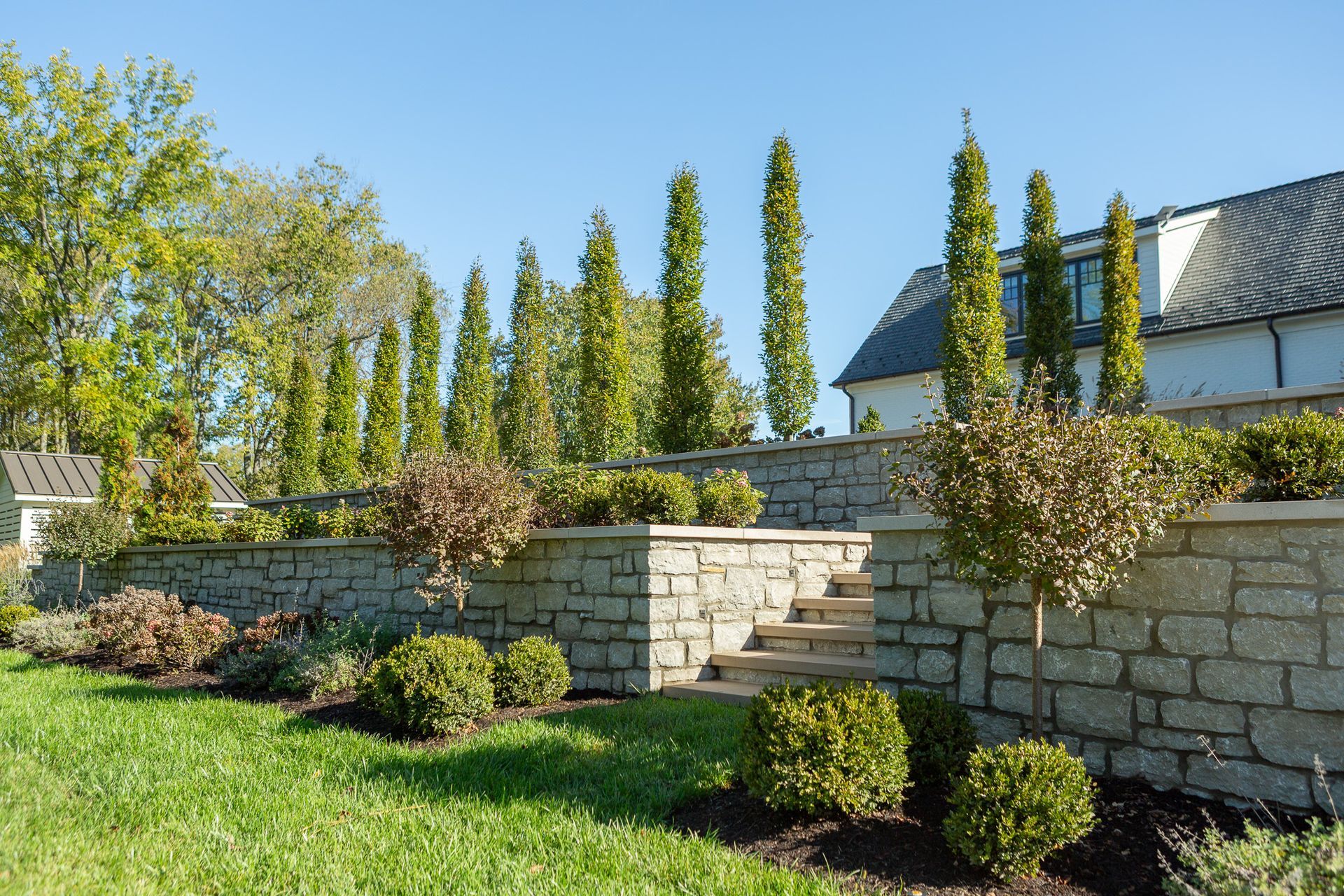 Stone retaining wall with steps, manicured landscaping, tall green trees, and a house under a blue sky.