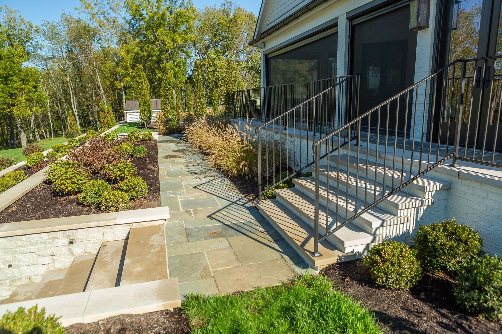Stone pathway leading to steps up to a porch with railings. Landscaping includes green shrubs and trees.
