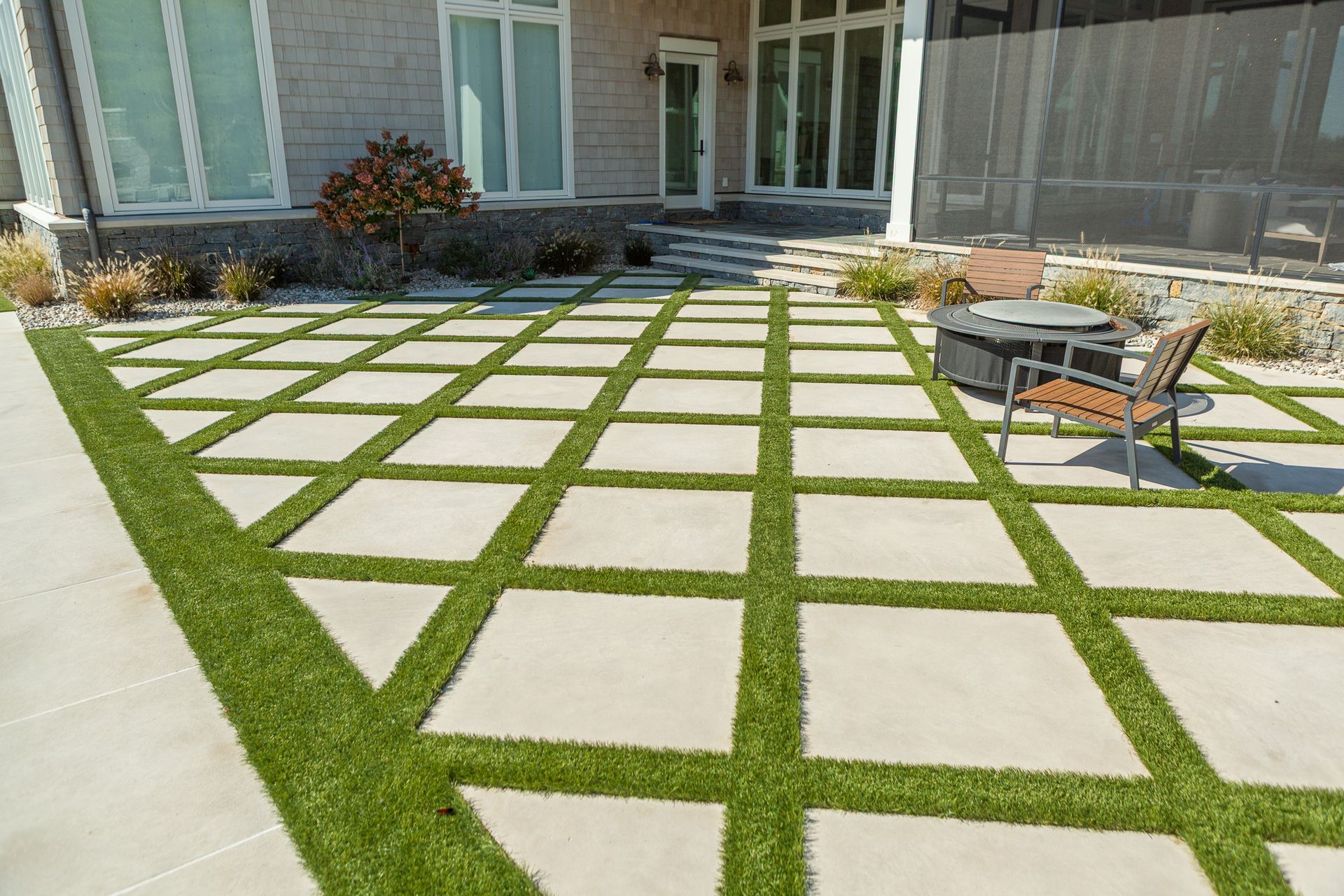 Patio with square concrete pavers and green artificial turf grid pattern. Fire pit and chairs.