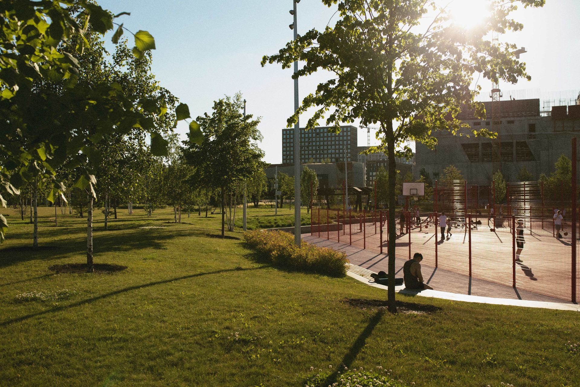 Park with green grass, trees, and a red sports area. A person sits on a bench. Buildings in the background.
