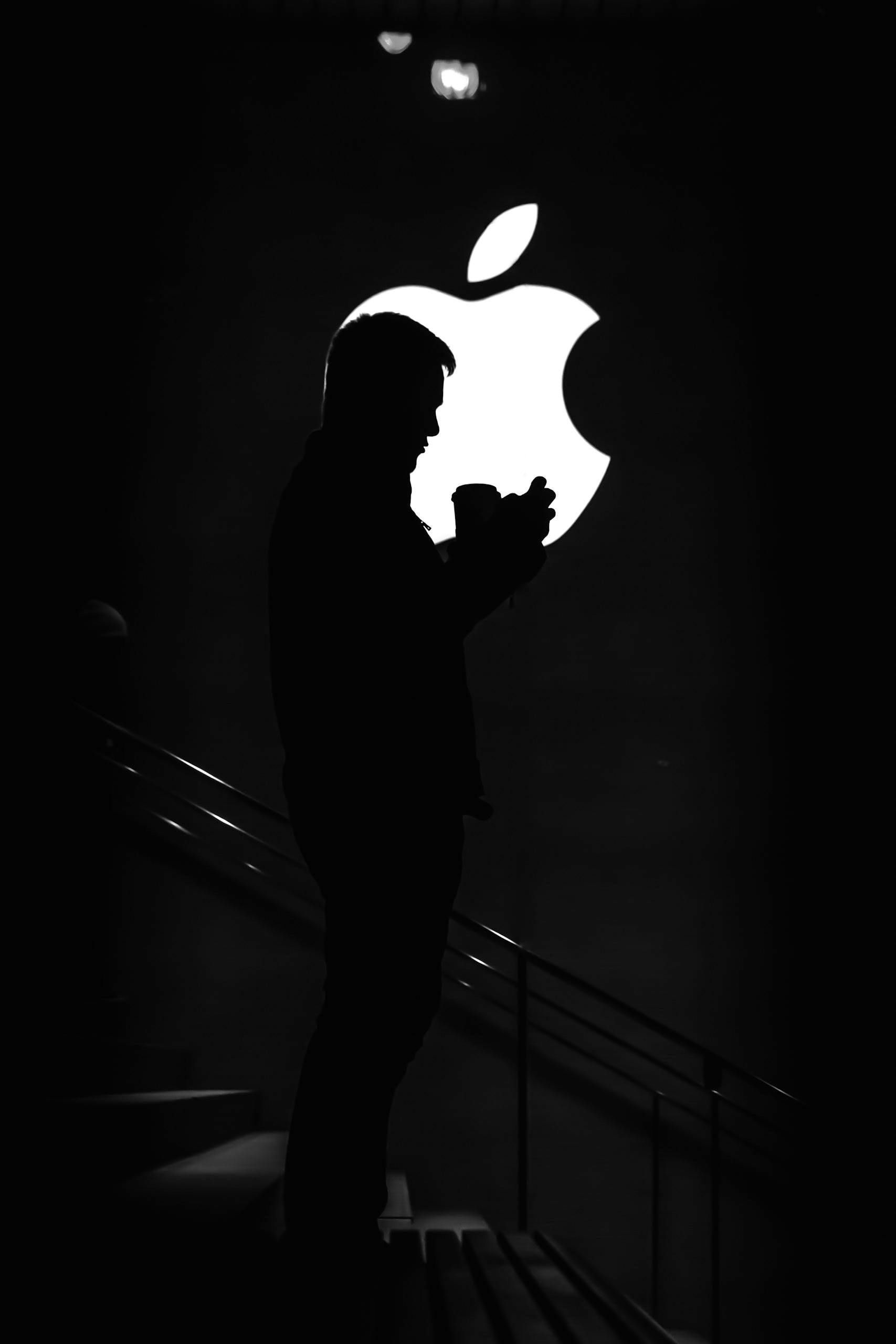 Silhouette of a person looking at their phone in front of a lit-up Apple logo in a dark store.