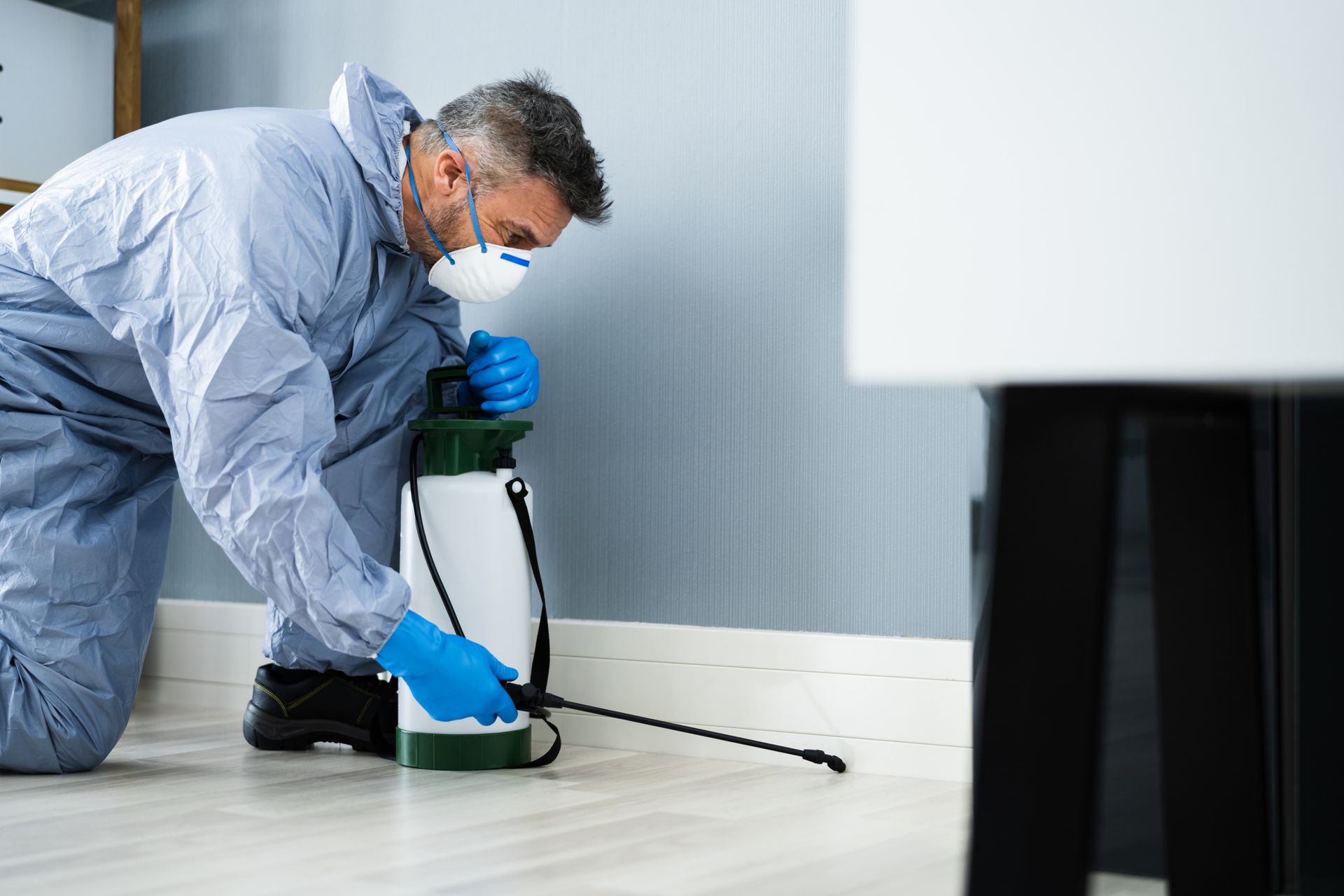 Male pest control exterminator in white safety gear sprays pesticide on the floor of a living room.