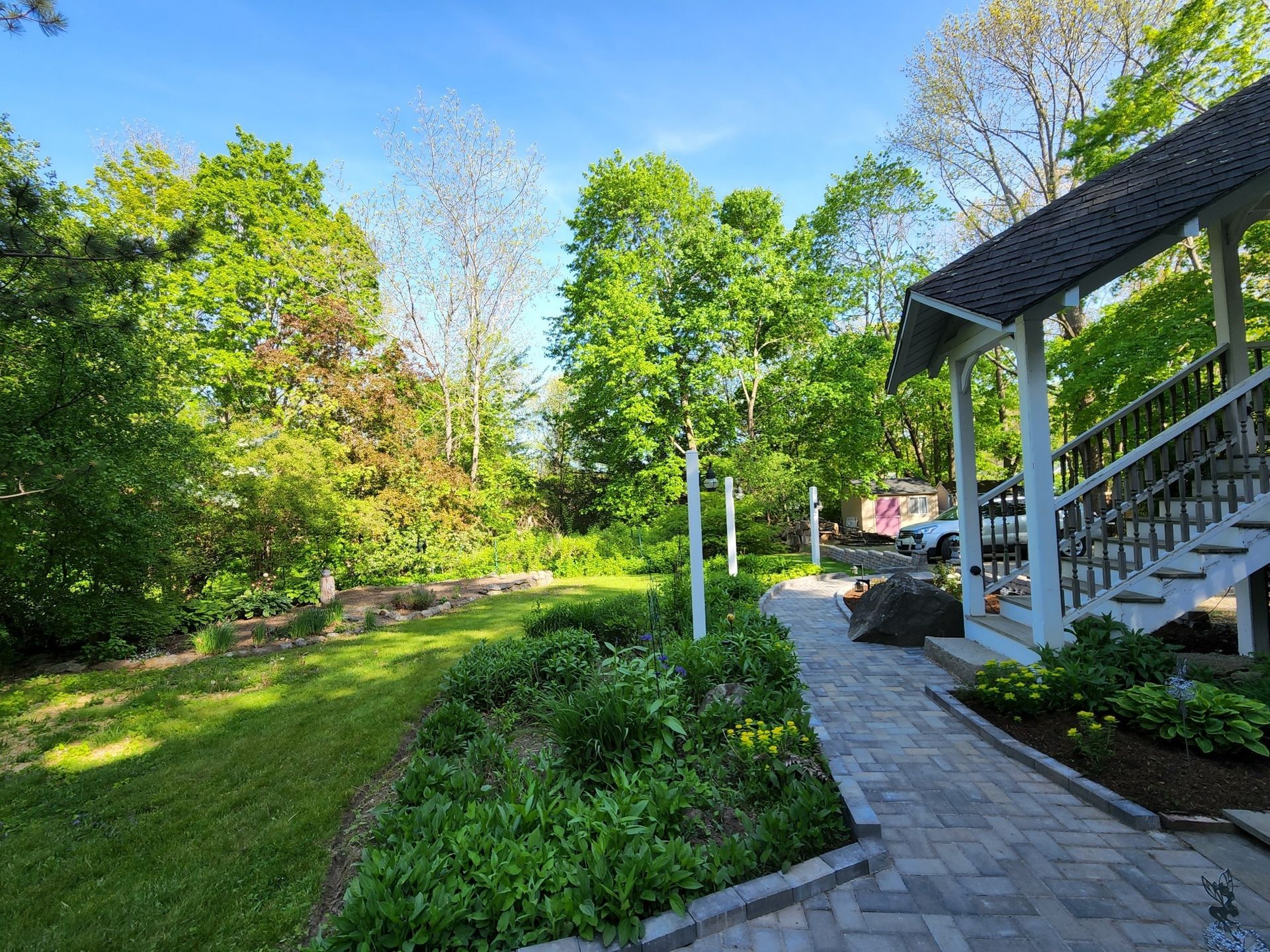 A brick walkway leading to a house with a porch and stairs surrounded by trees.