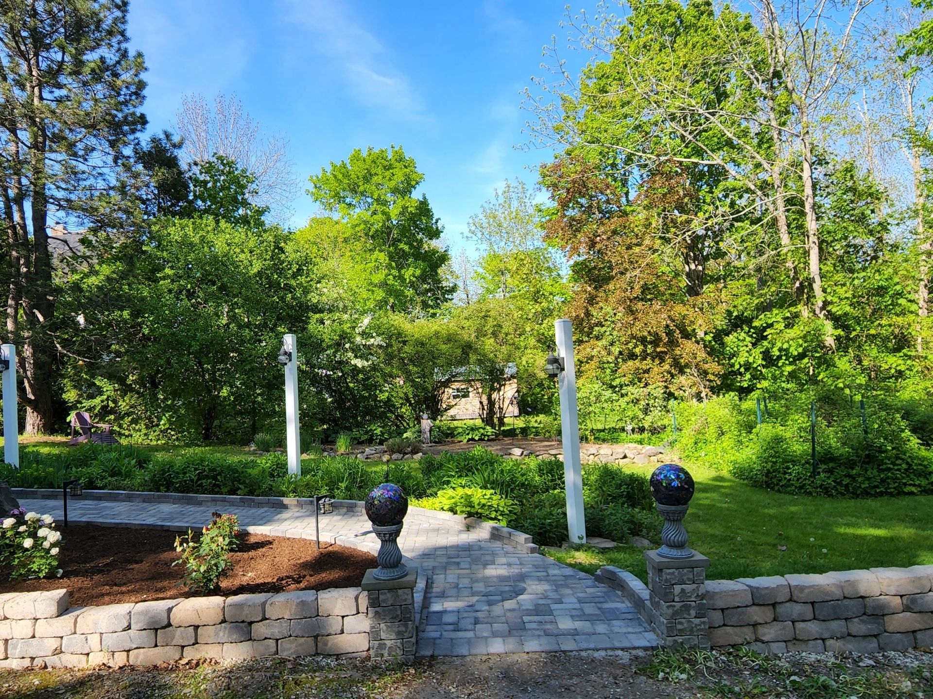 A brick walkway leading to a garden surrounded by trees on a sunny day.