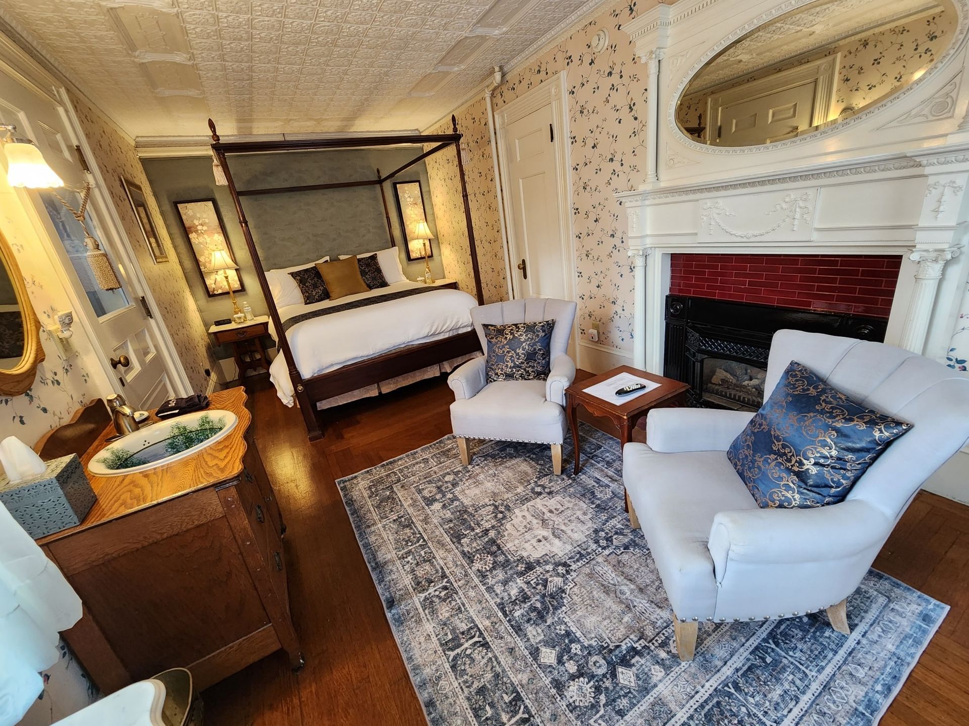 A bedroom with a four poster bed , chair , dresser and fireplace.