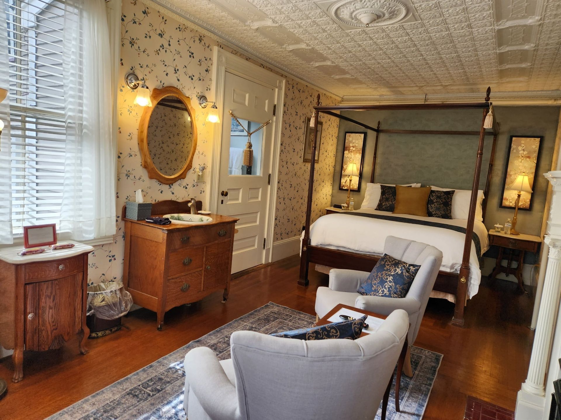 A bedroom with a four poster bed , chairs , dresser and mirror.