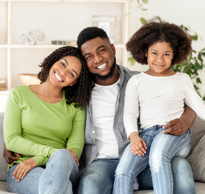 Family smiling together on a couch; woman in green, man in gray, child in white, bright room.