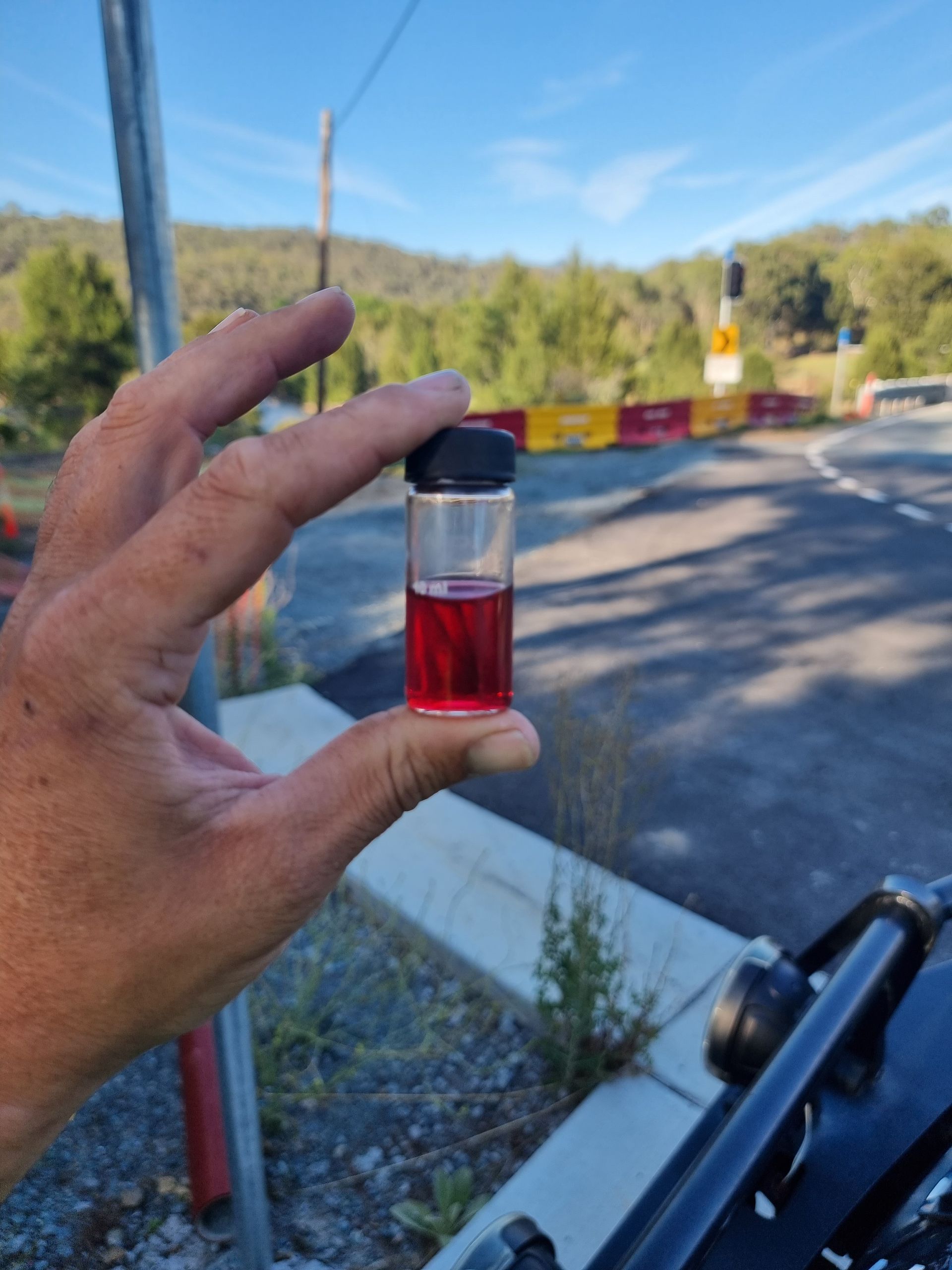 Hand holding a vial of red liquid, roadside setting with a blurred background of trees and road — Pro Pipe Solutions in Queanbeyan, NSW