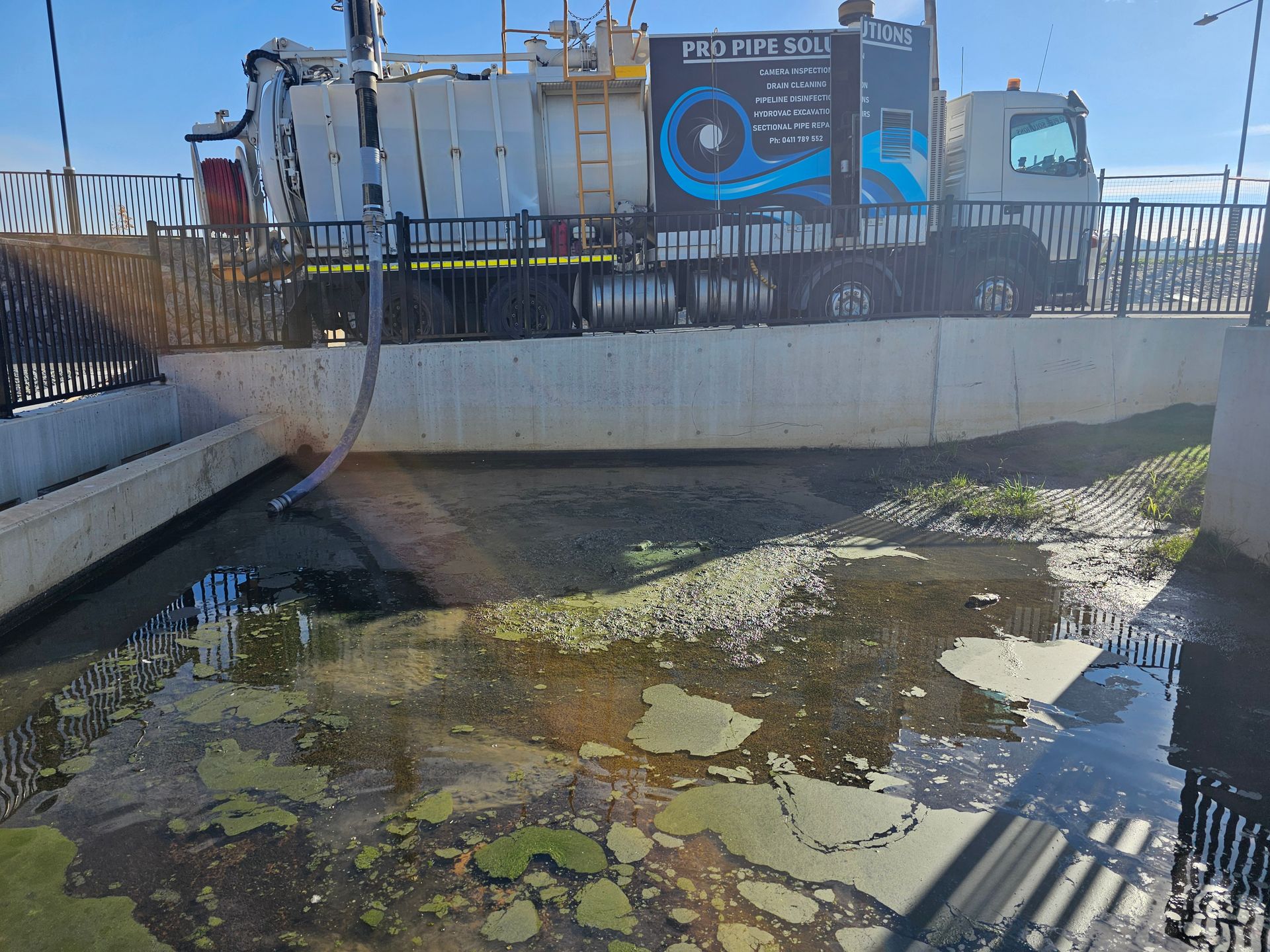 A truck cleaning a polluted waterway. Gray concrete and metal fencing surrounds the water with algae — Pro Pipe Solutions in Queanbeyan, NSW