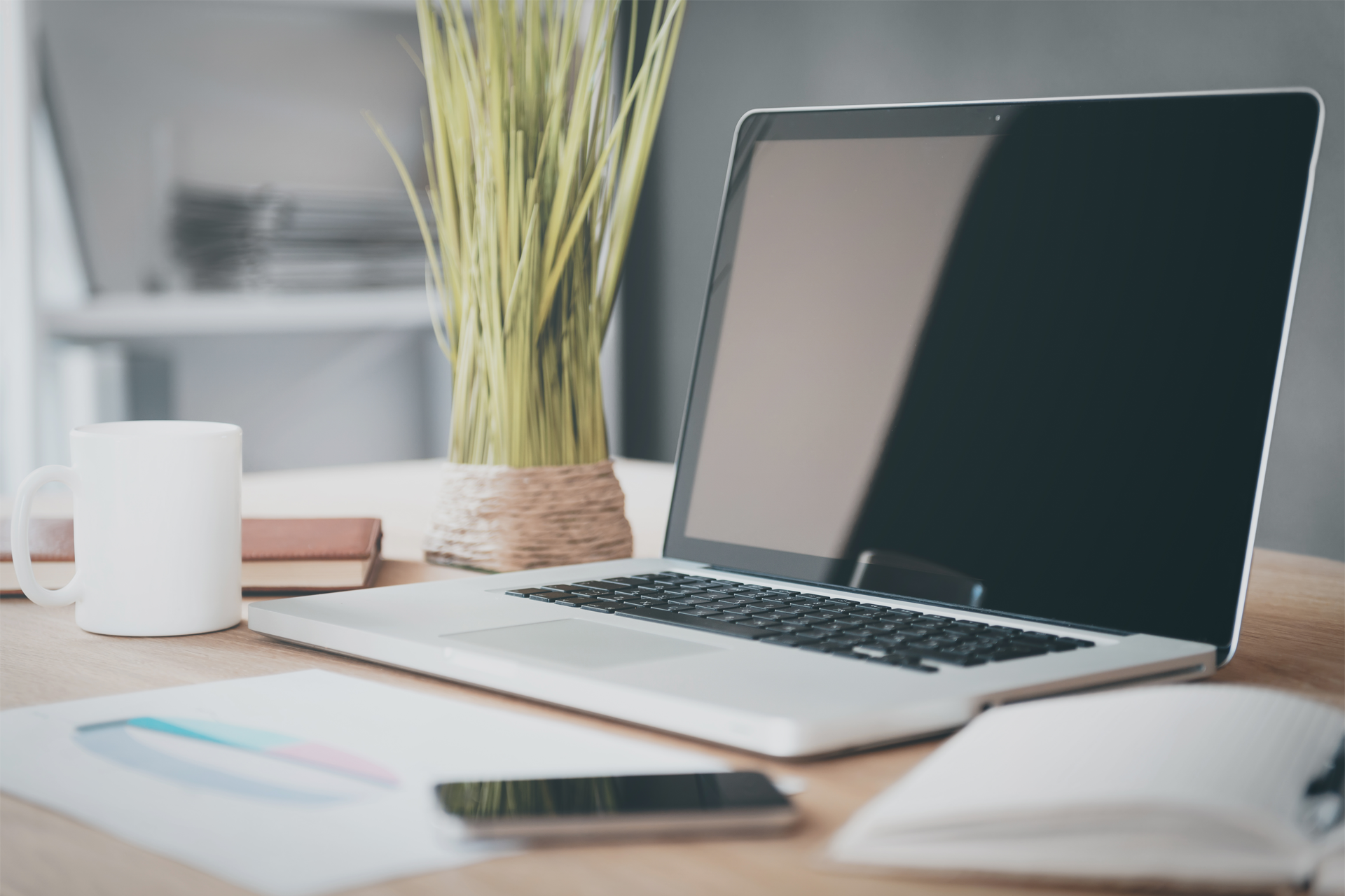 A laptop computer is sitting on a desk next to a vase of grass.