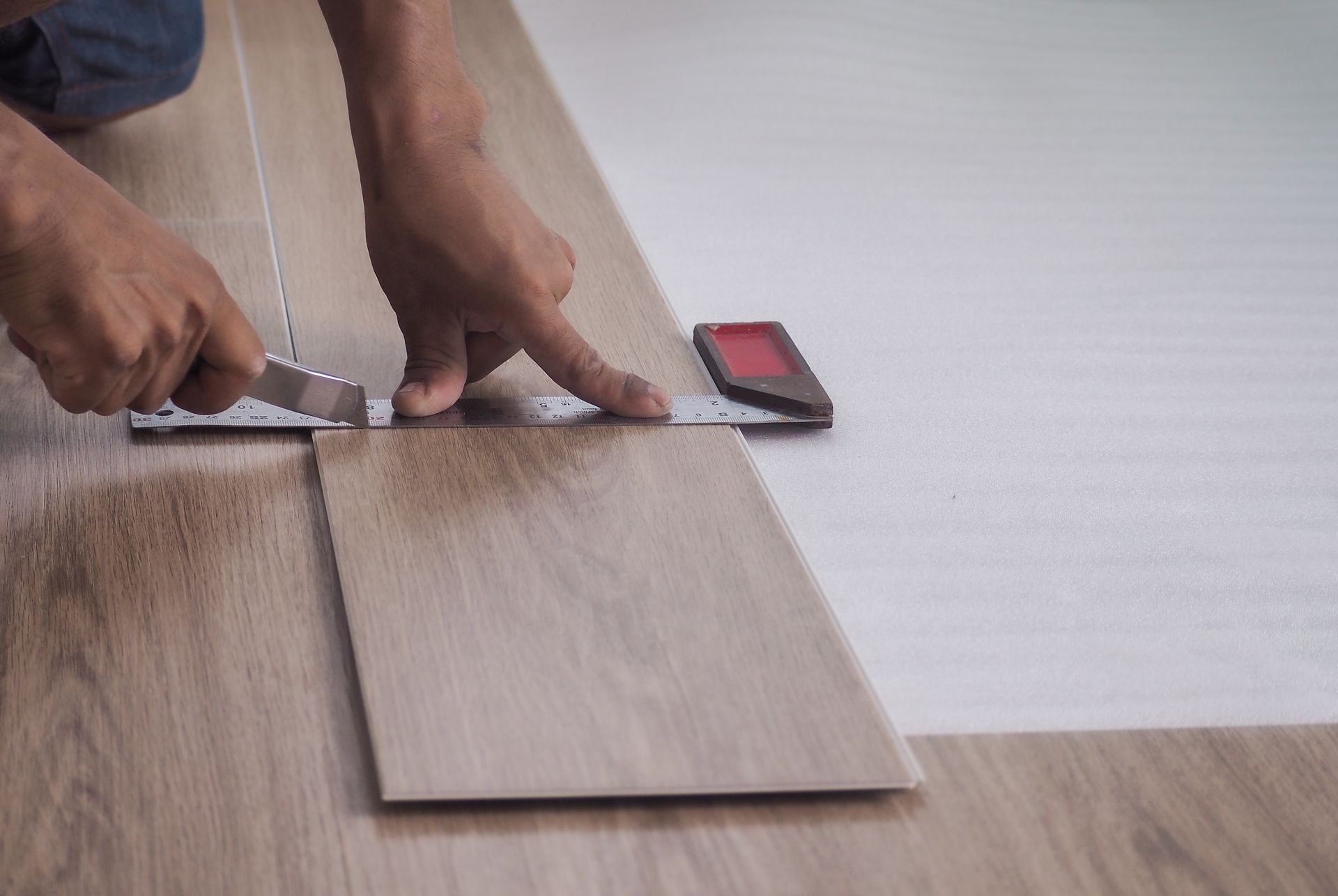 Person cutting flooring with a utility knife and a ruler in a room with light-colored floors.