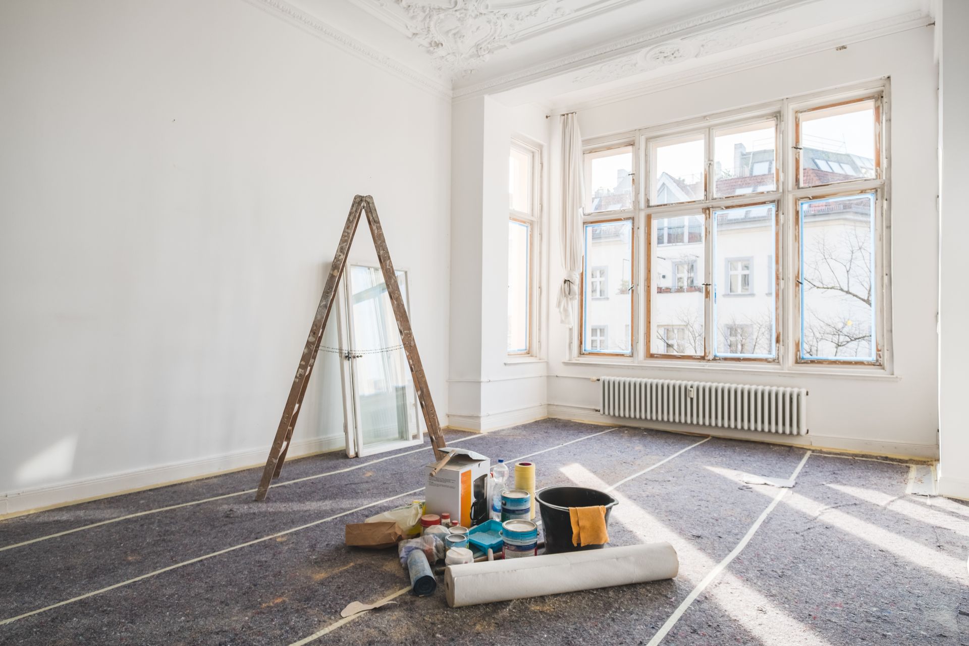 Empty room undergoing renovation with a ladder, tools, and window overlooking buildings. White walls and gray floor covering.