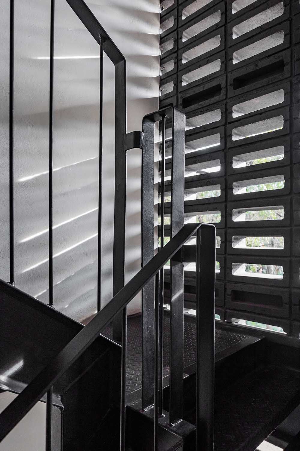Escalera y barandilla de metal negro junto a una pared de ladrillo oscuro y texturizado; la luz del sol proyecta sombras.