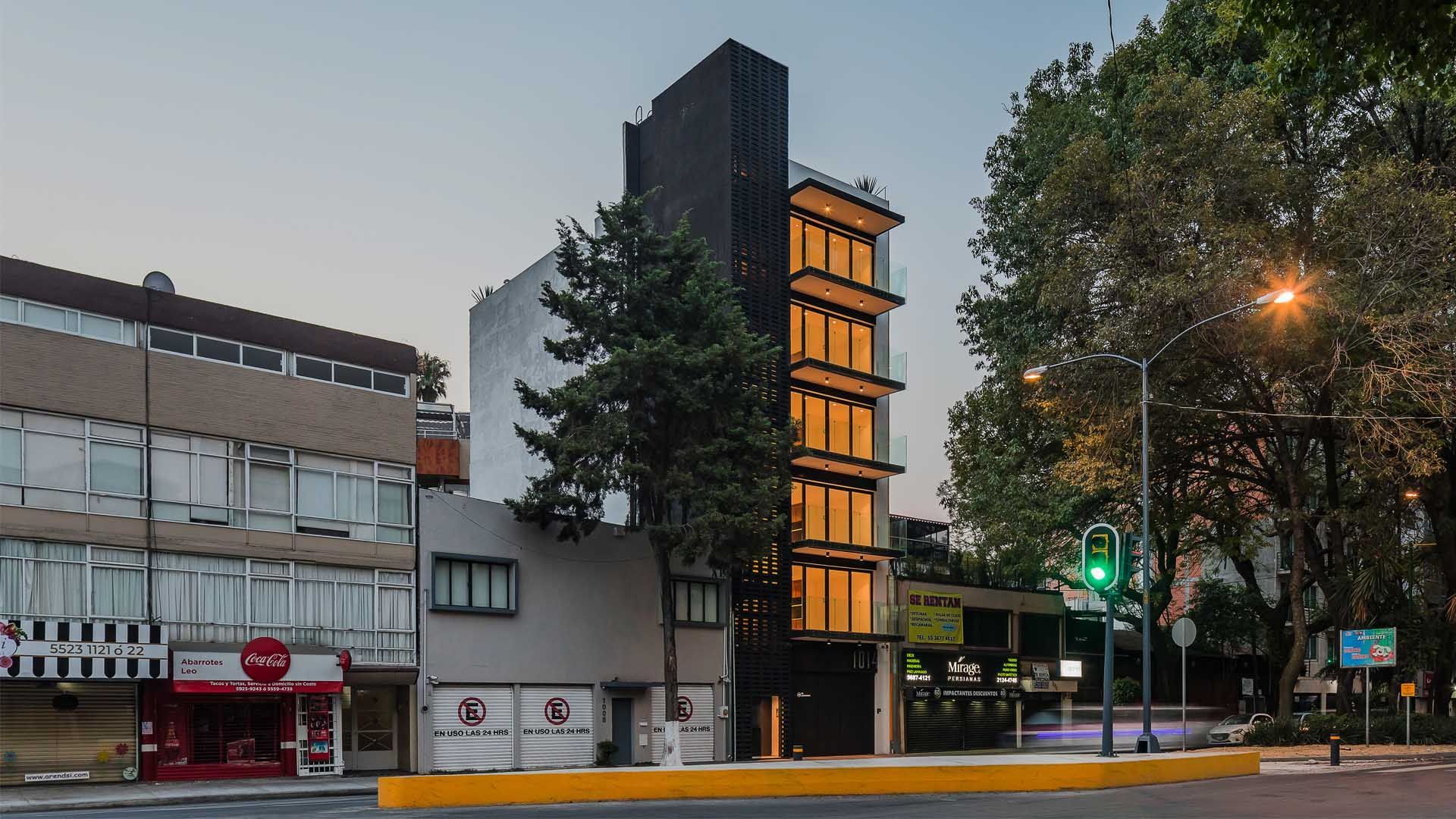 Edificio de varios pisos con una fachada vertical oscura al lado de un edificio blanco y gris; un árbol se encuentra al frente.