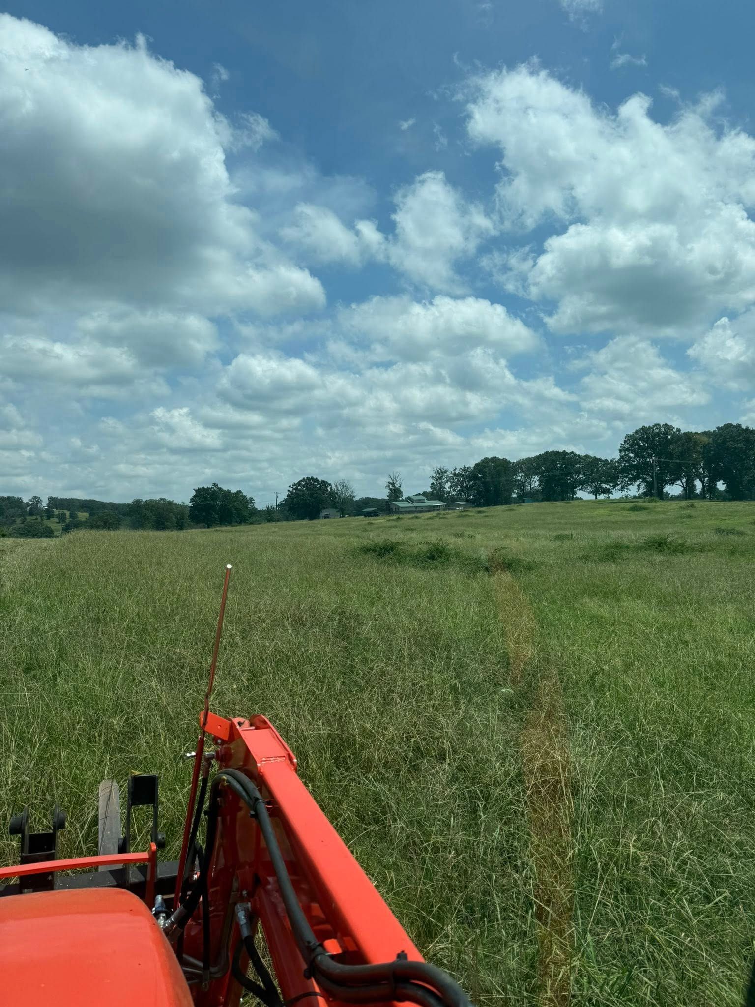 View from a tractor cab, looking out over a vast, green field under a bright, partly cloudy blue sky.