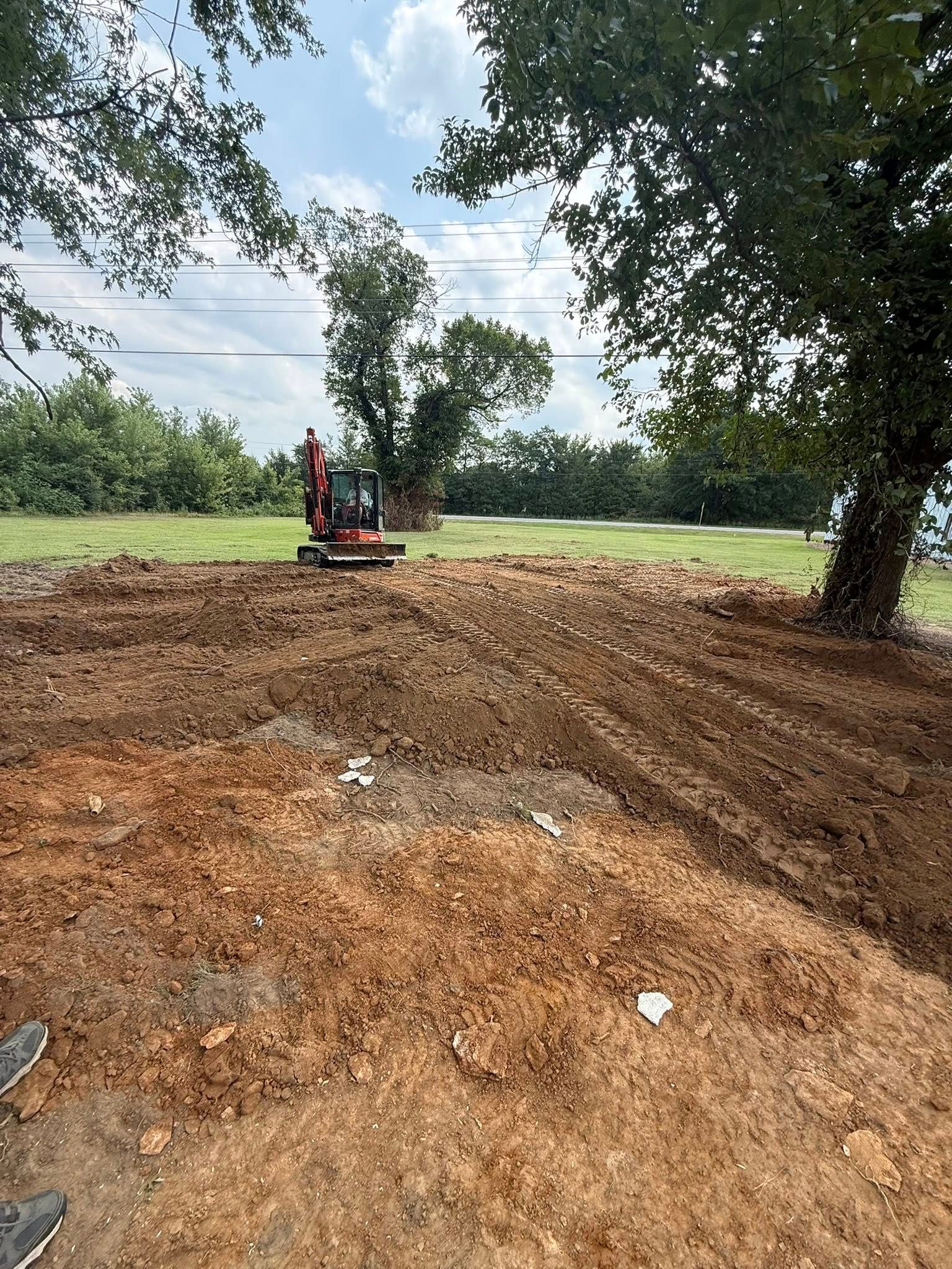 A red mini-excavator sits on a cleared, dirt-covered lot surrounded by trees under a partly cloudy sky.