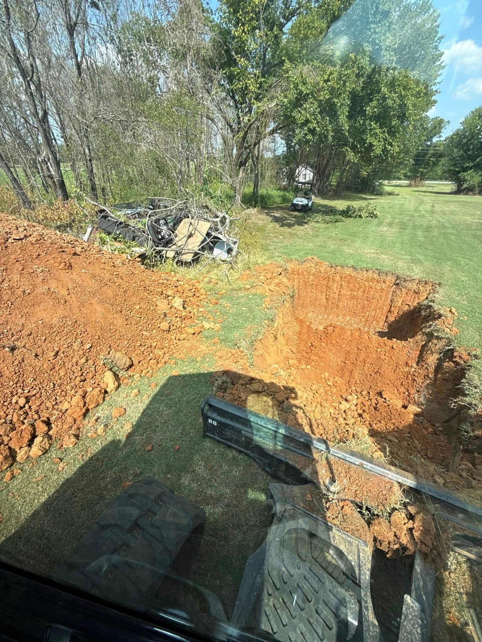 A view from an excavator cab showing a freshly dug trench and a pile of red dirt in a grassy field with trees.