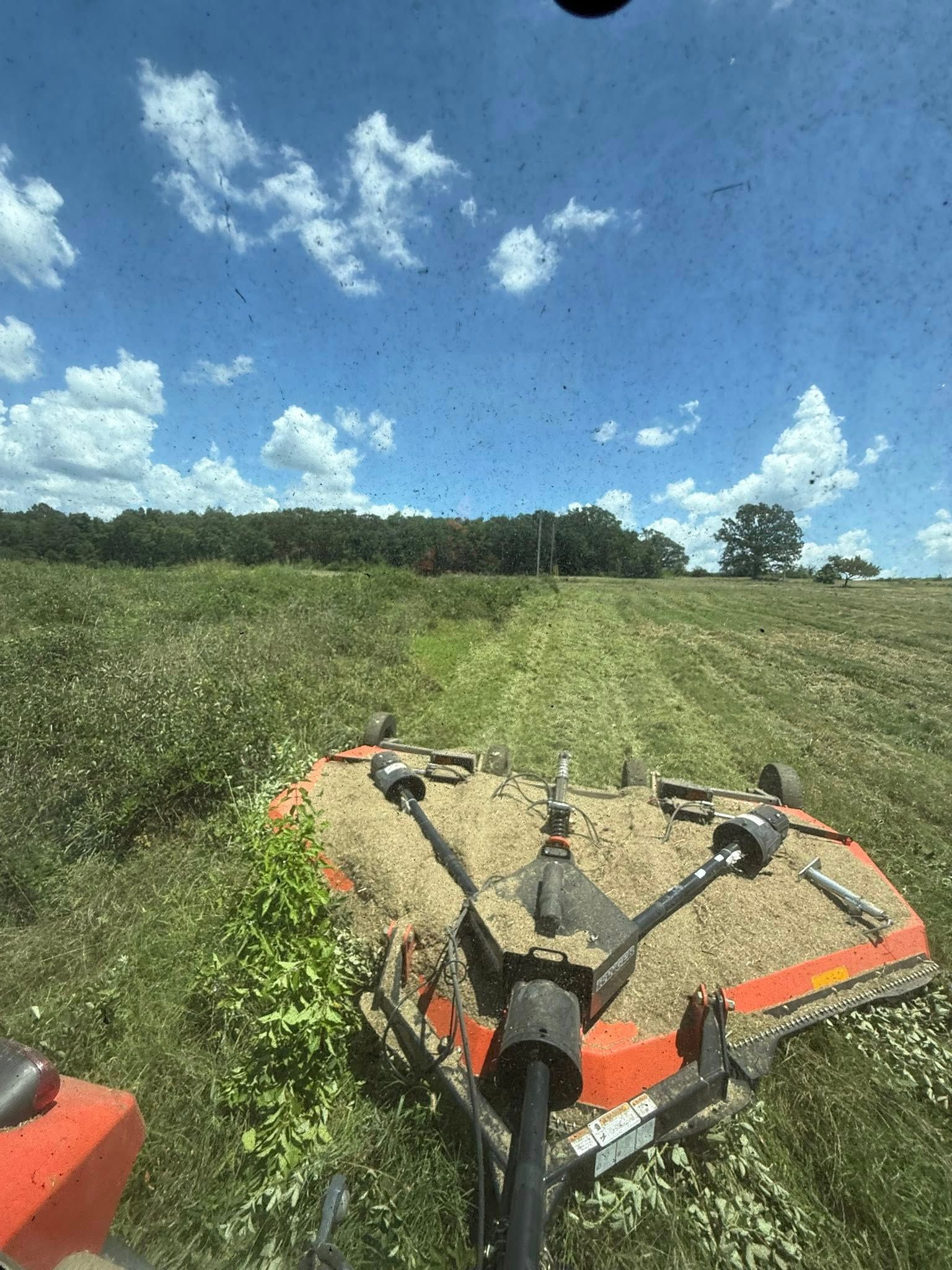 A view from a tractor cab looking down at an orange rotary cutter mowing tall grass in a sunny field.