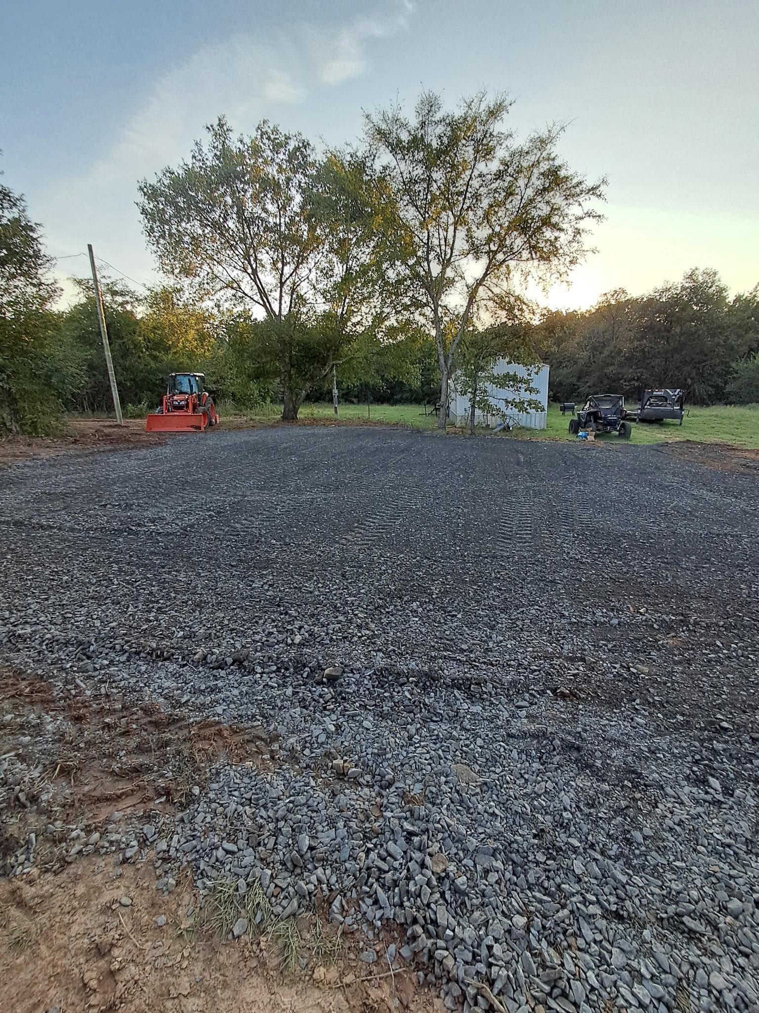 A gravel-covered construction site in a grassy field with trees in the background and heavy machinery parked nearby.