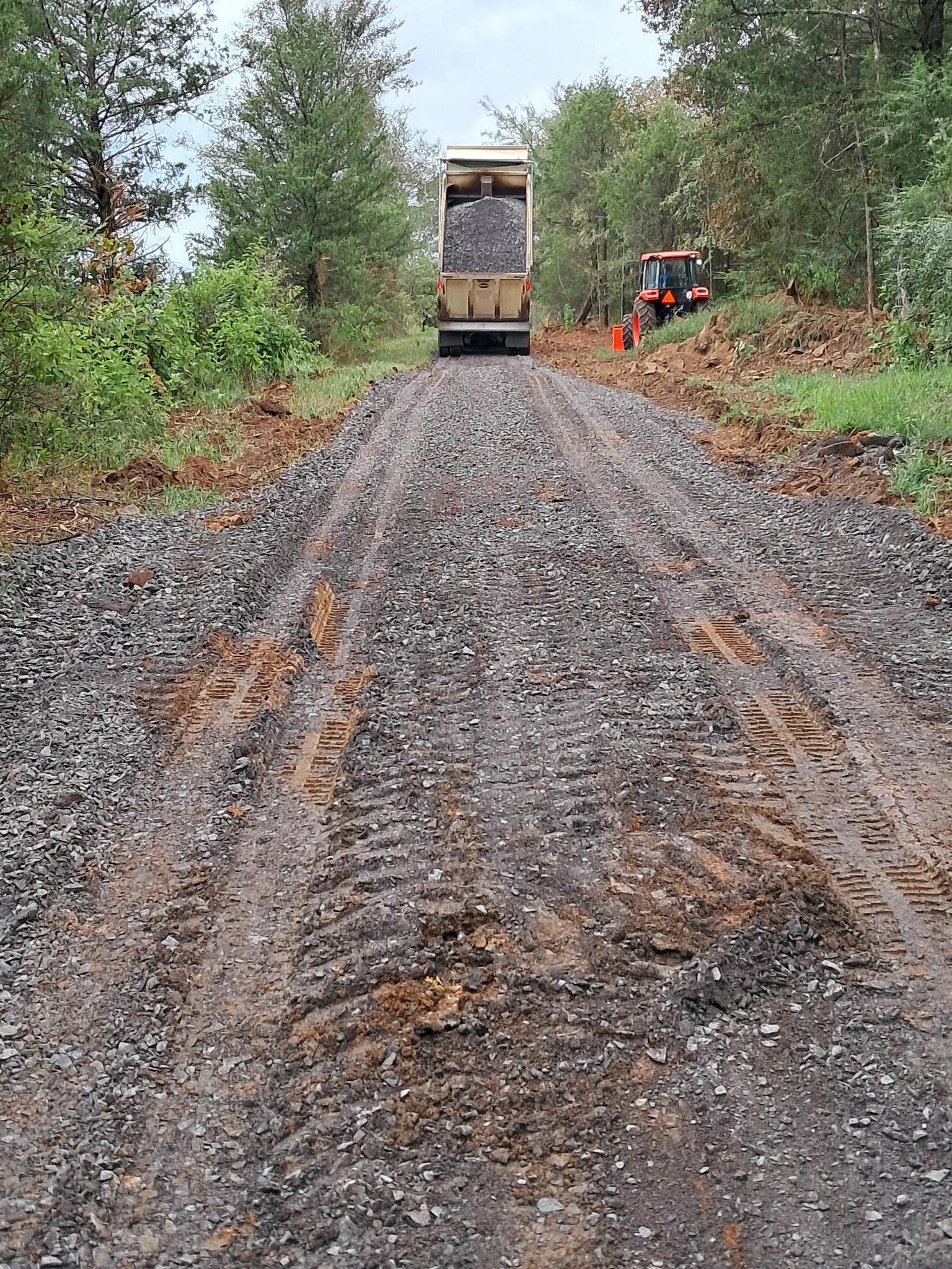 A dump truck spreads gravel on a muddy, wooded road while a small tractor follows alongside.