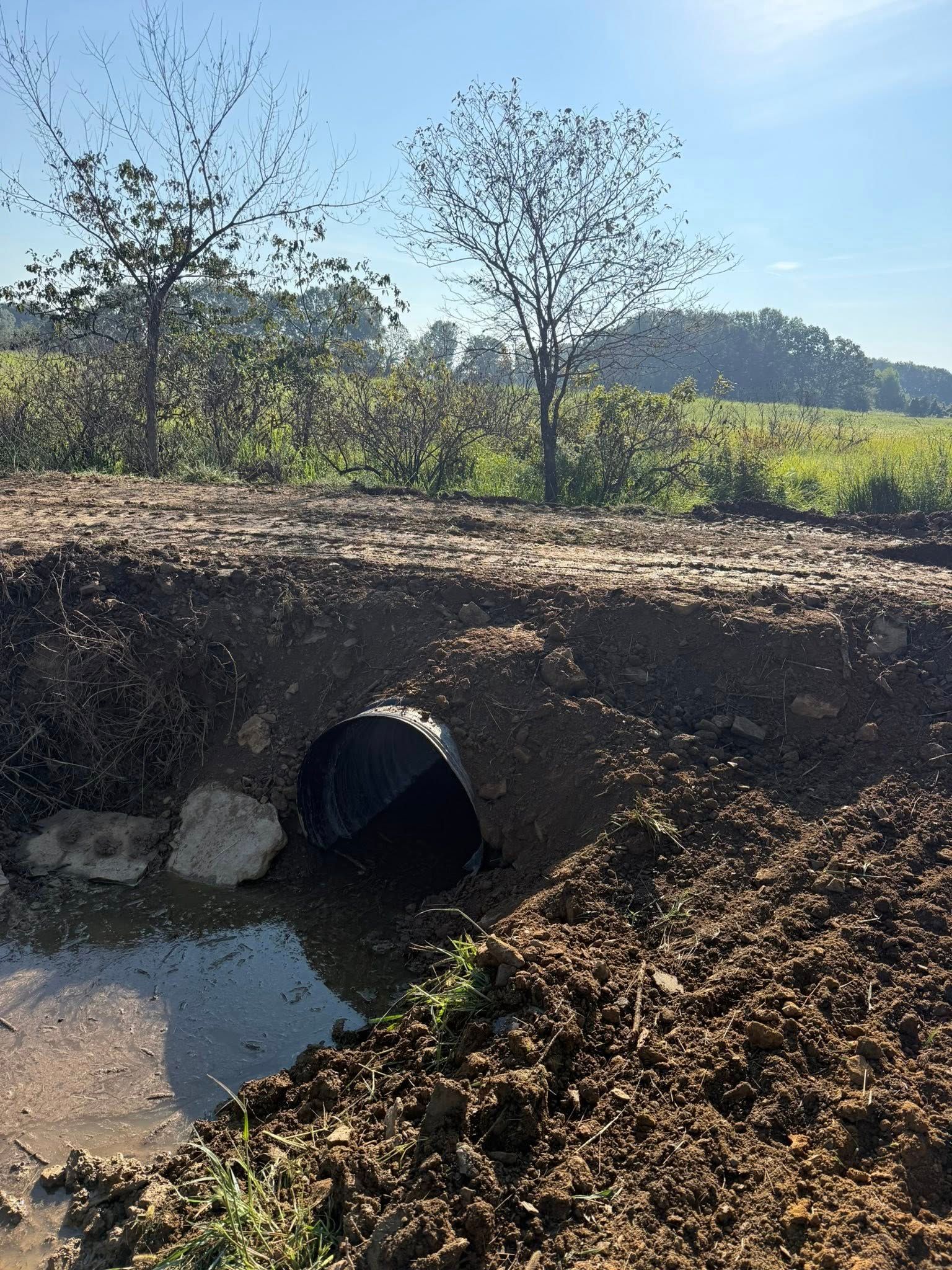 A black culvert pipe sits in a muddy, excavated trench near a rural landscape with trees under a blue sky.