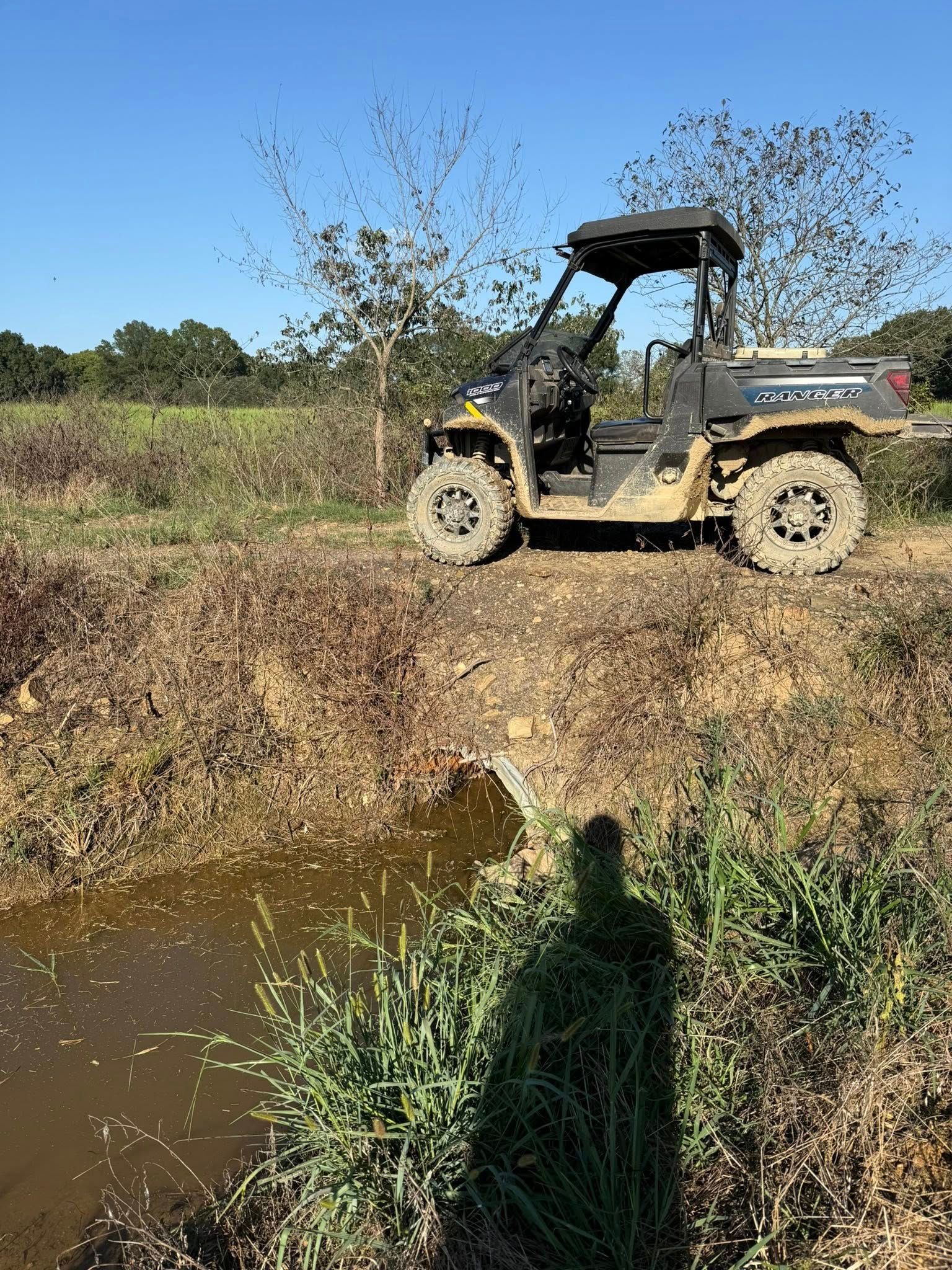 A grey utility vehicle (UTV) parked on a dirt embankment next to a small, muddy creek under a clear blue sky.