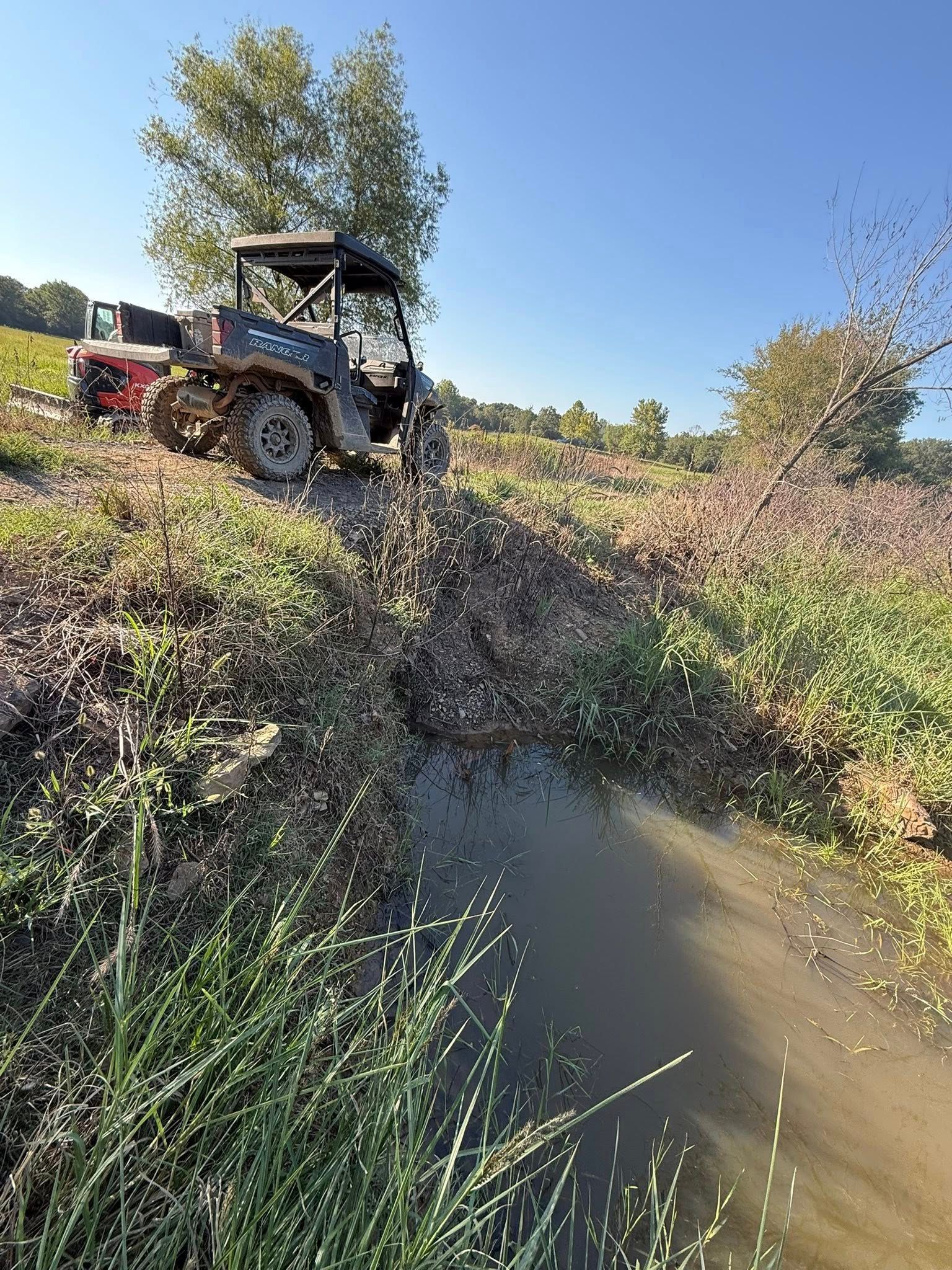 A dark utility terrain vehicle (UTV) parked on a grassy embankment beside a muddy creek under a clear blue sky.
