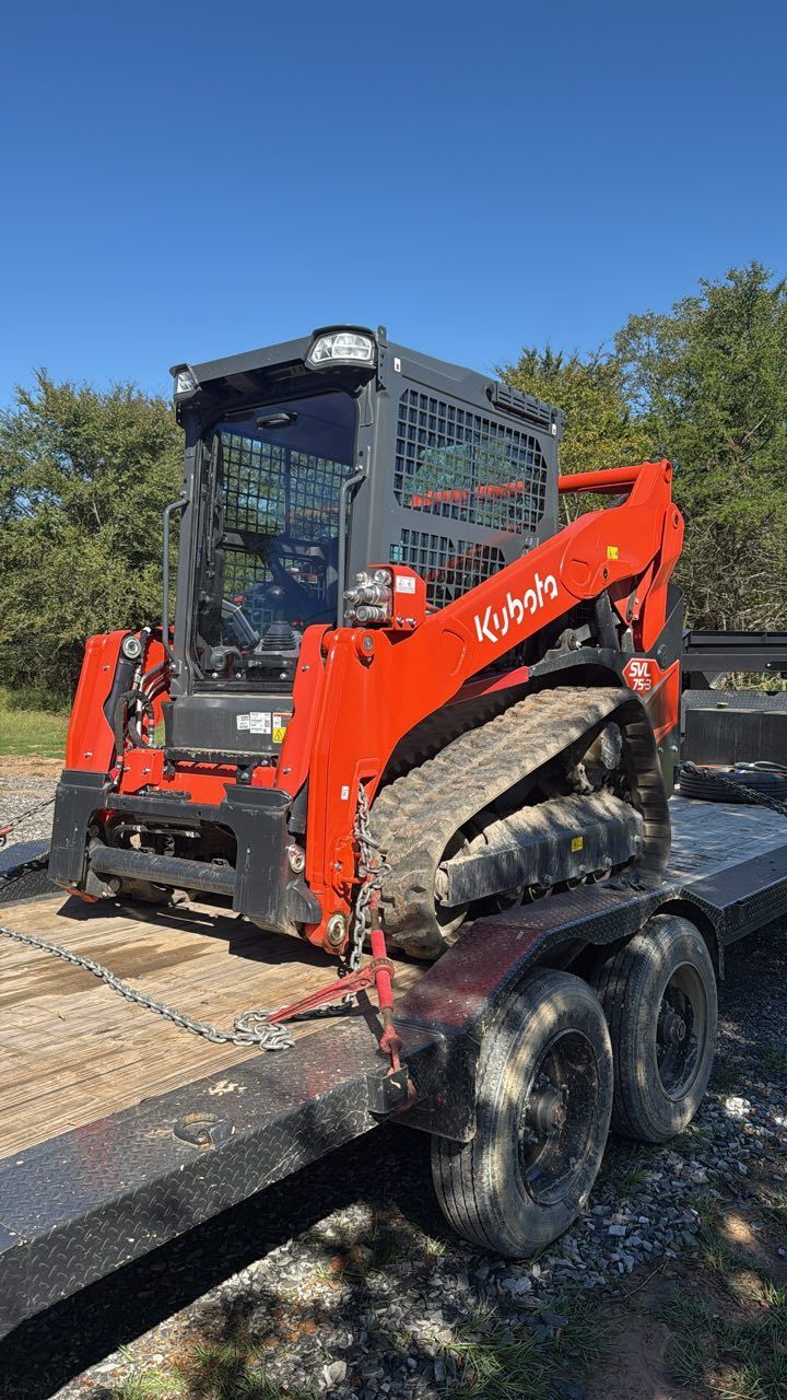 An orange Kubota compact track loader secured to a flatbed trailer outdoors under a clear blue sky.