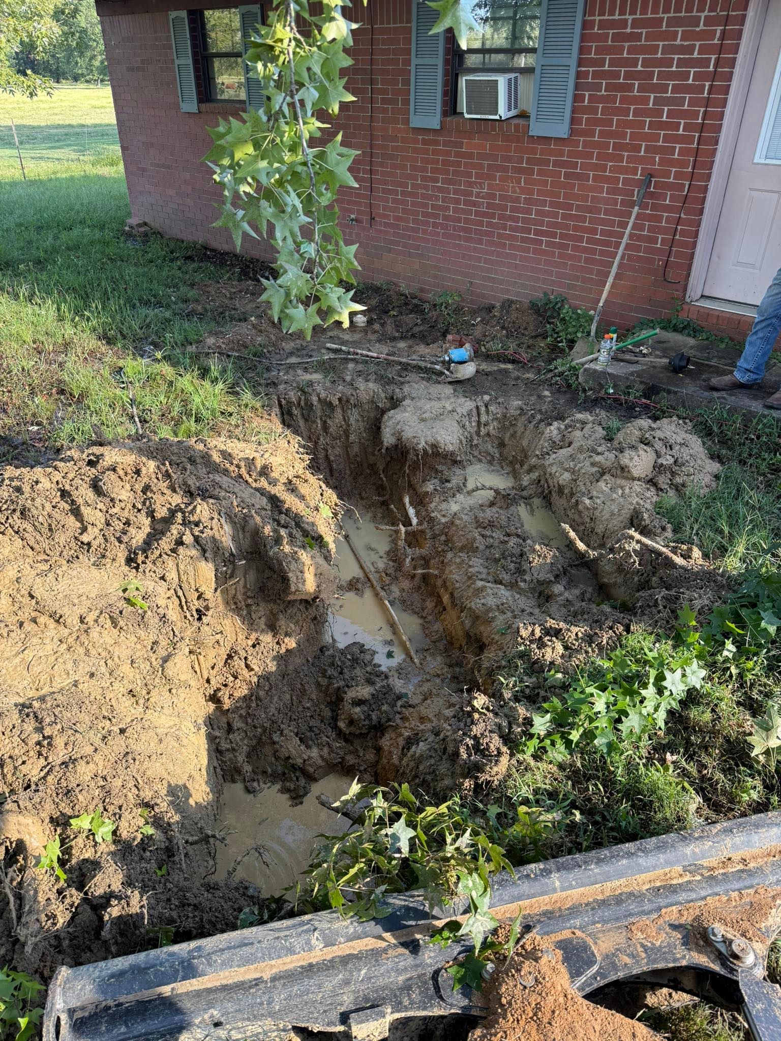 A muddy, open trench dug in the lawn alongside a red brick house, with a segment of pipe visible at the bottom.