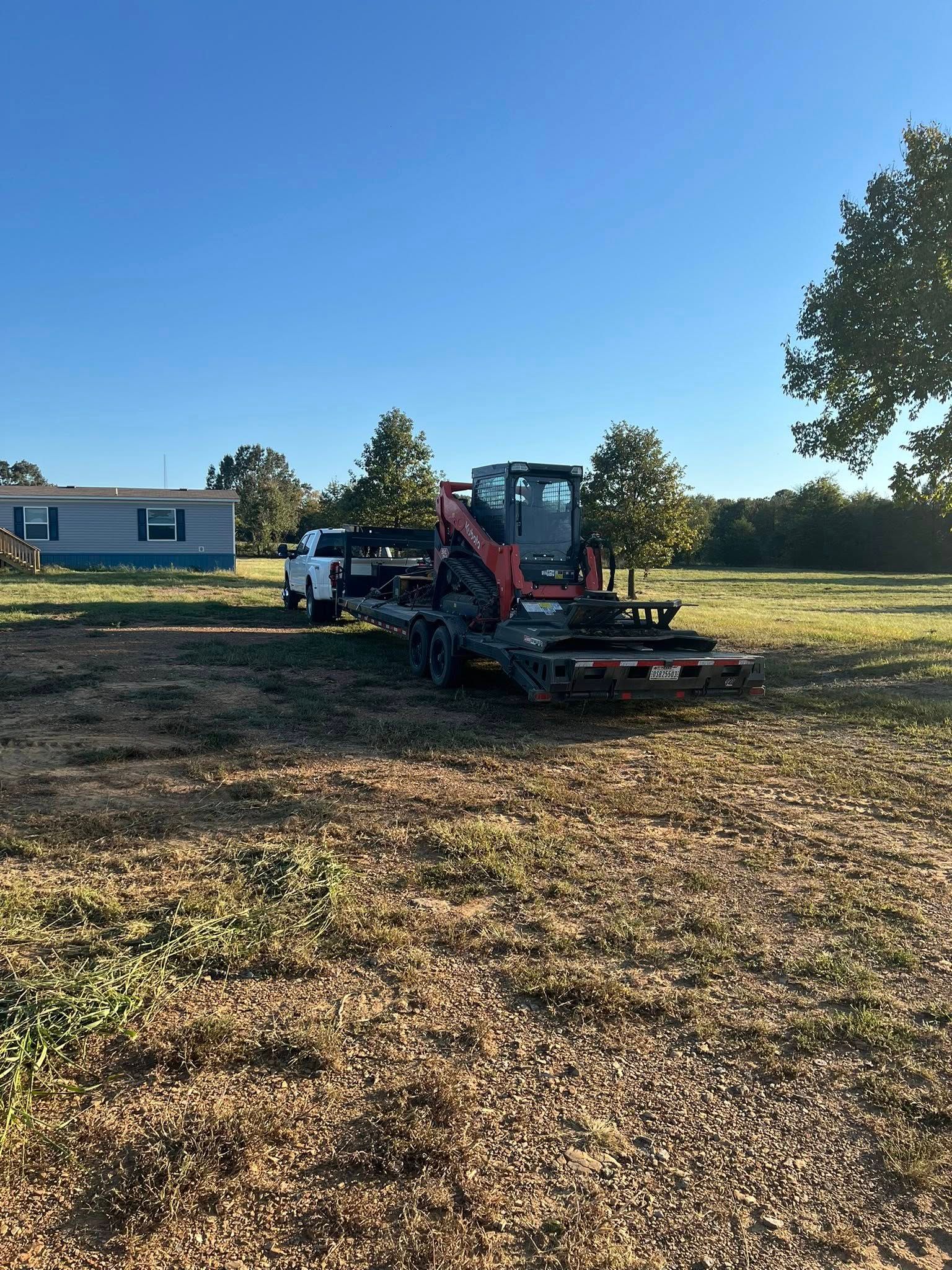 An orange skid steer loader is loaded onto a flatbed trailer attached to a white pickup truck in a grassy field.