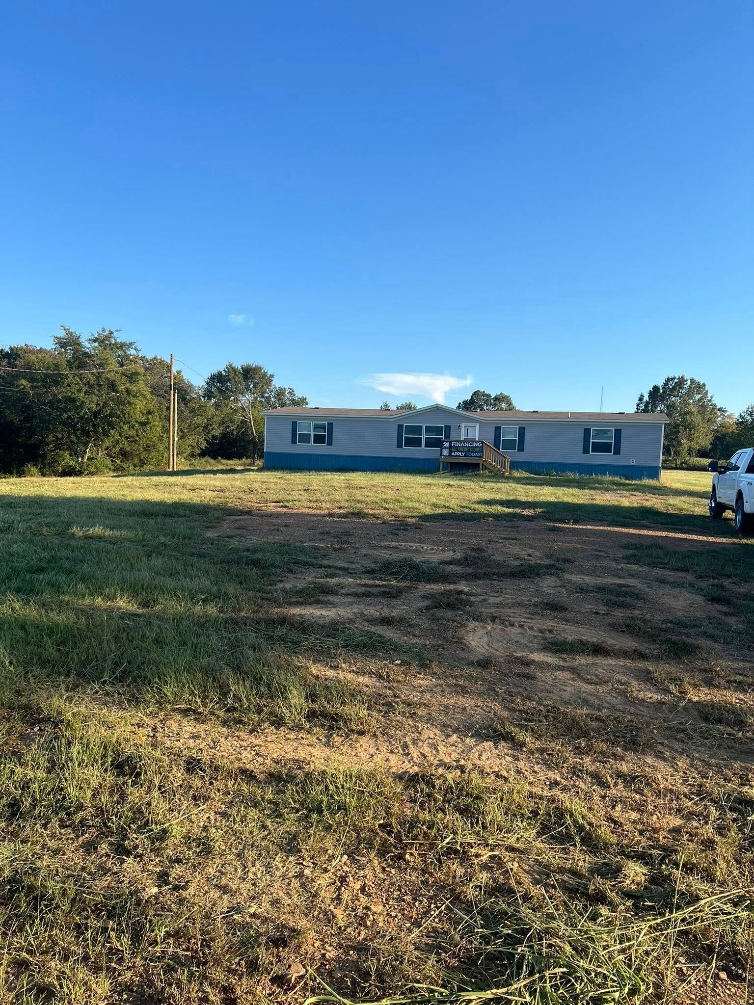 A light-colored mobile home sits in the background of a large, grassy lot under a clear blue sky.
