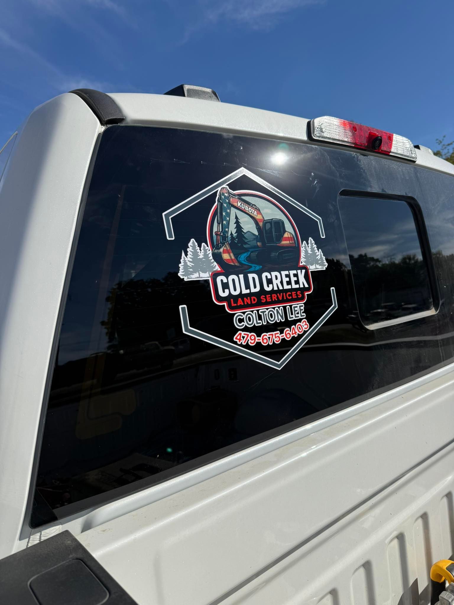 A decal on the rear window of a white truck showing an excavator inside a hexagon with the text Cold Creek Landscaping.