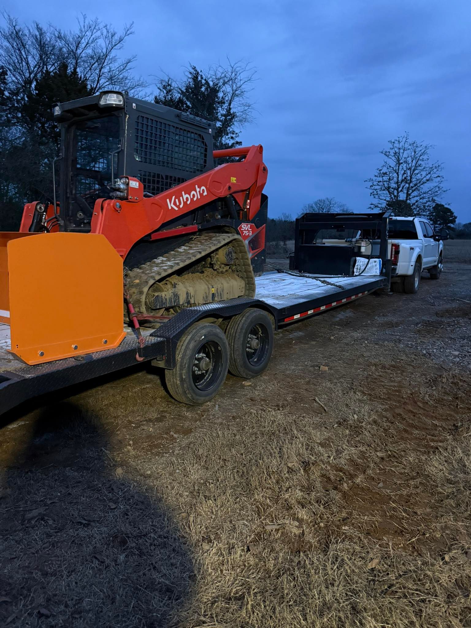 A bright orange compact track loader sits on a flatbed trailer attached to a white pickup truck in a grassy field.
